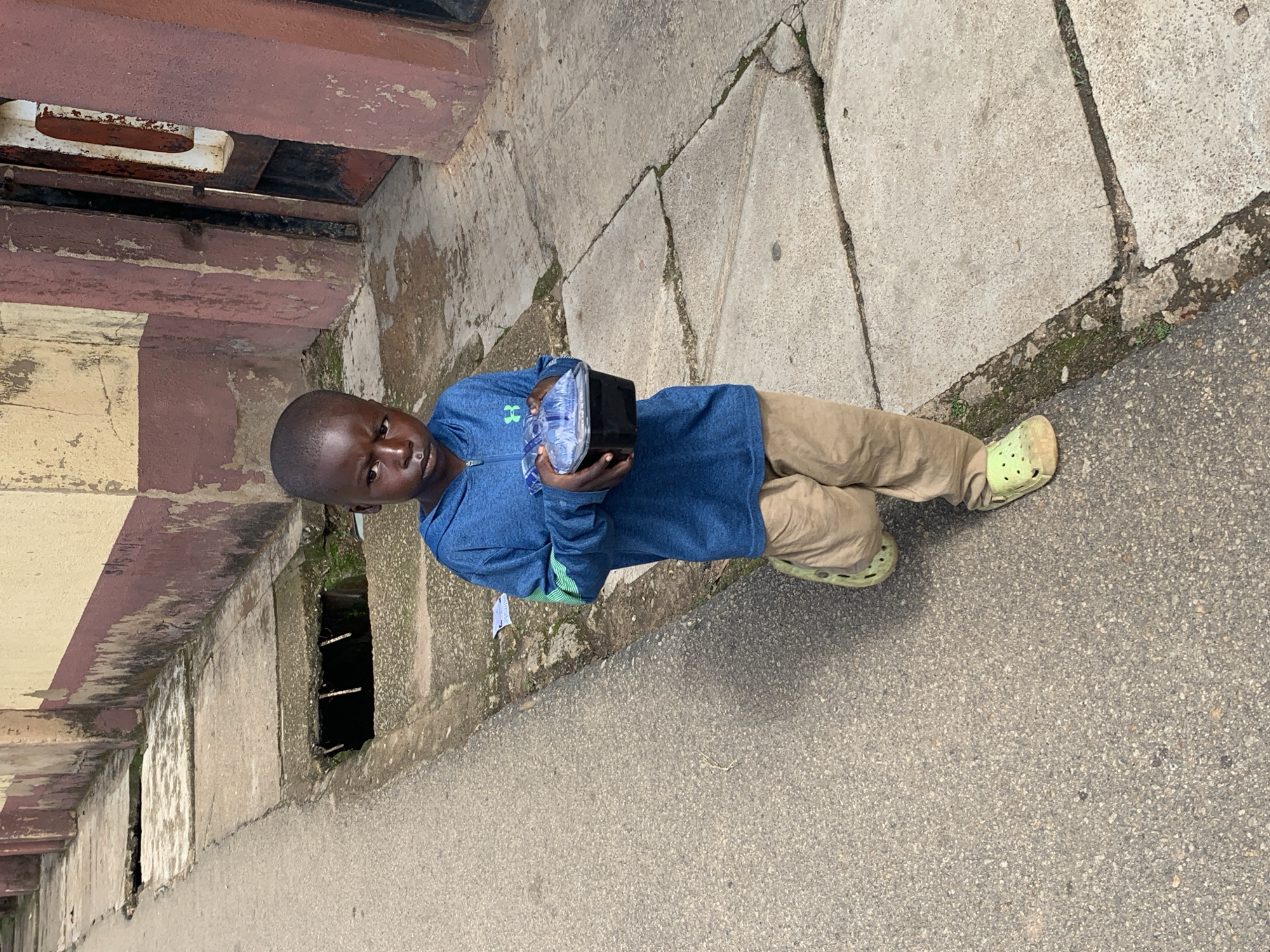 In Jos, Nigeria, a young boy is captured mid-stride on a concrete sidewalk. He is wearing a blue hooded sweatshirt with a green stripe detail on the sleeves, tan pants, and lime green clogs. In his hands, he holds a black plastic container covered with a clear plastic lid. The boy looks directly at the camera with a serious expression. The background consists of a building with a textured, weathered facade painted in shades of beige and maroon. A metal grate is visible in the concrete drainage along the sidewalk. The scene appears to be a street in an urban setting, likely a residential or commercial area in Jos.