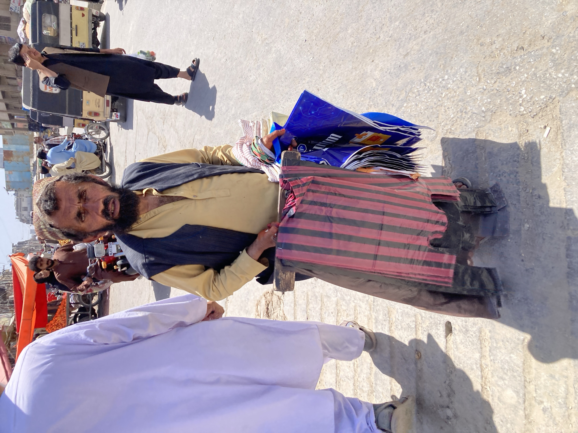 The image shows a middle-aged man in Quetta City Tehsil, Pakistan, standing outdoors on a bright day. He has a dark beard and is wearing a light yellow shirt, a dark vest, and sandals. He is carrying a variety of items, including colorful plastic bags and striped cloth, likely for sale. The setting appears to be a street or market area, with other people, vehicles (including a rickshaw), and market stalls visible in the background. A person wearing a white robe is partially visible in the foreground. The ground is paved with concrete.