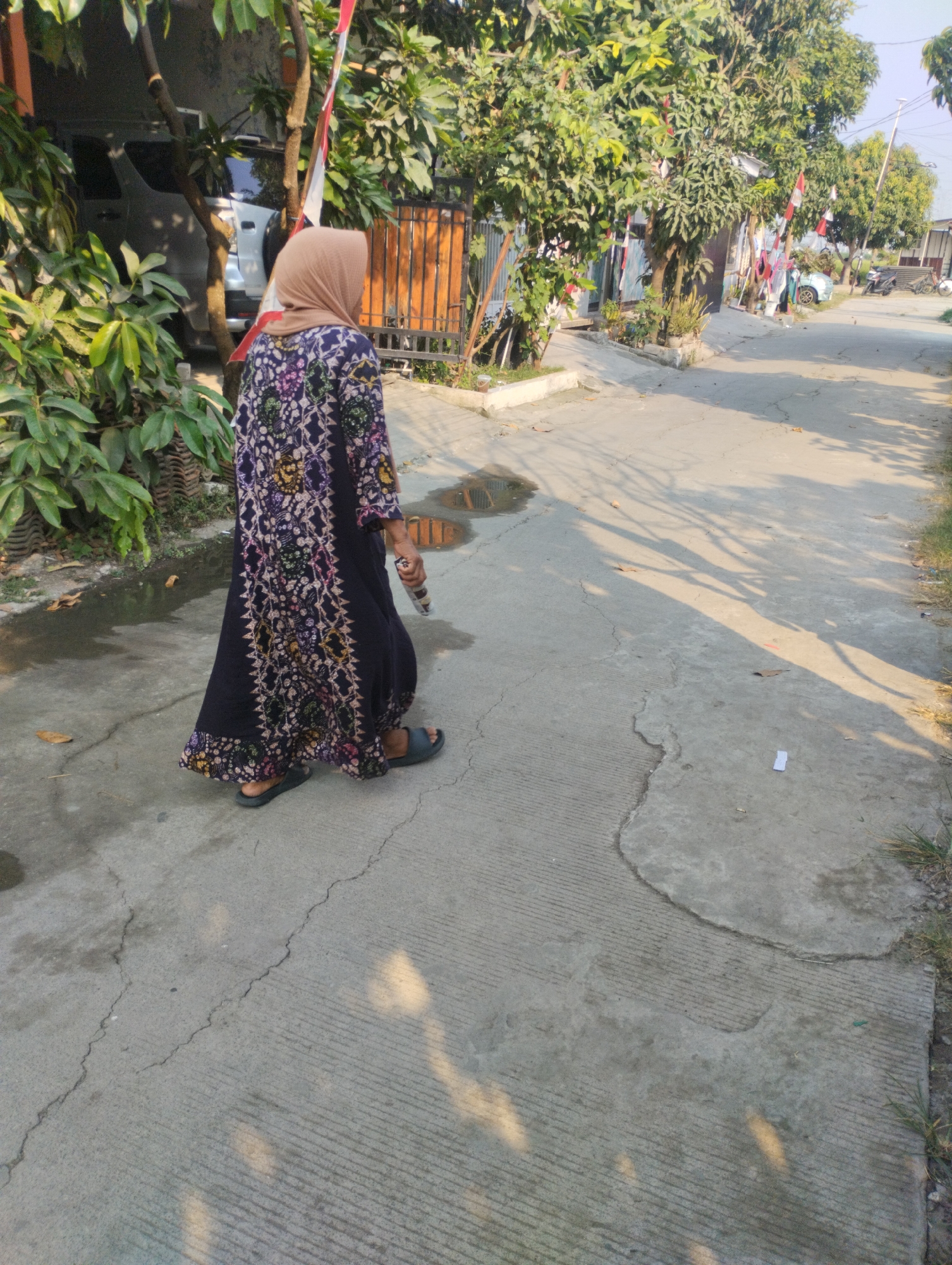 The image depicts a street scene in Karangraharja, Indonesia, under bright daylight. A person, viewed from the rear, is walking on a concrete pathway. They are wearing a long, dark-colored dress with a patterned print in various colors, including purple, pink, and yellow. A light brown hijab covers their head. The person is wearing dark open-toed sandals. They are holding a cylindrical object in their right hand.
The environment includes a street or pathway with visible cracks and some areas with standing water. To the left of the pathway, there is lush green vegetation and trees. In the background, there are houses with fences and parked vehicles, including a silver SUV partially visible. Several Indonesian flags are displayed, attached to poles near the houses and trees, indicating a possible celebration or national holiday. The shadows of trees are cast across the concrete surface, suggesting the sun is positioned at an angle. The overall scene suggests a residential area in a tropical climate.