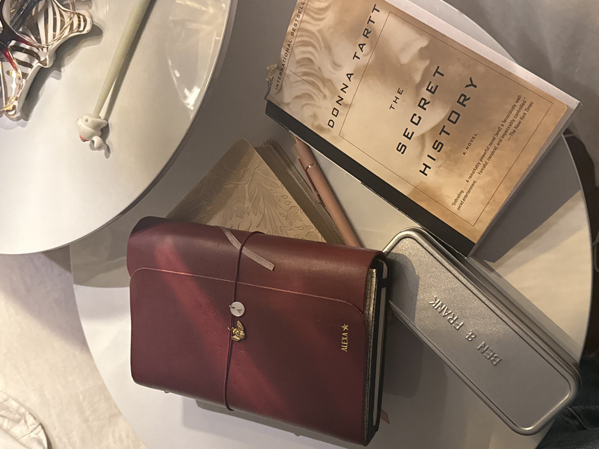 Here is a forensic description of the image:
**Scene:** The image presents a close-up, eye-level shot of a table arrangement.
**Subjects/Objects:** Prominent items include:
- A copy of Donna Tartt's novel "The Secret History".
- A red leather-bound journal or notebook with a charm and the name "ALEXA" imprinted.
- A metal container labeled "BEN & FRANK".
- A pen.
- Several other small objects and decorative items.
**Environment:** The objects are arranged on a white, multi-tiered table. The background is indistinct but appears to be an indoor setting.
There is no indication of the city of Mexico City, Mexico in the image.
