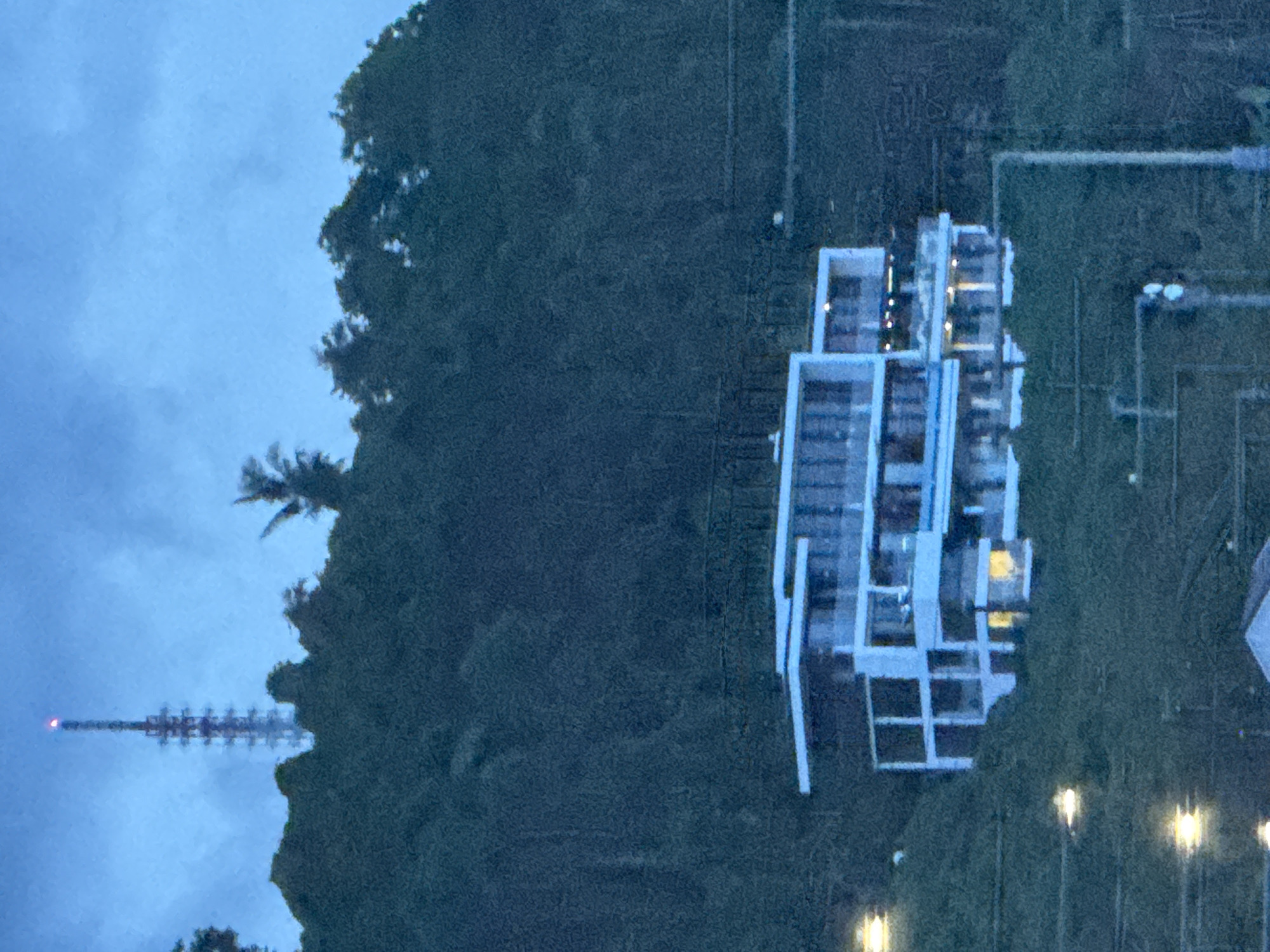 The image shows an aerial view of a building with a white frame structure located in Davao City, Philippines. The building is situated near a heavily wooded area. A communications tower is visible in the distance. Streetlights illuminate the foreground. The sky is overcast.