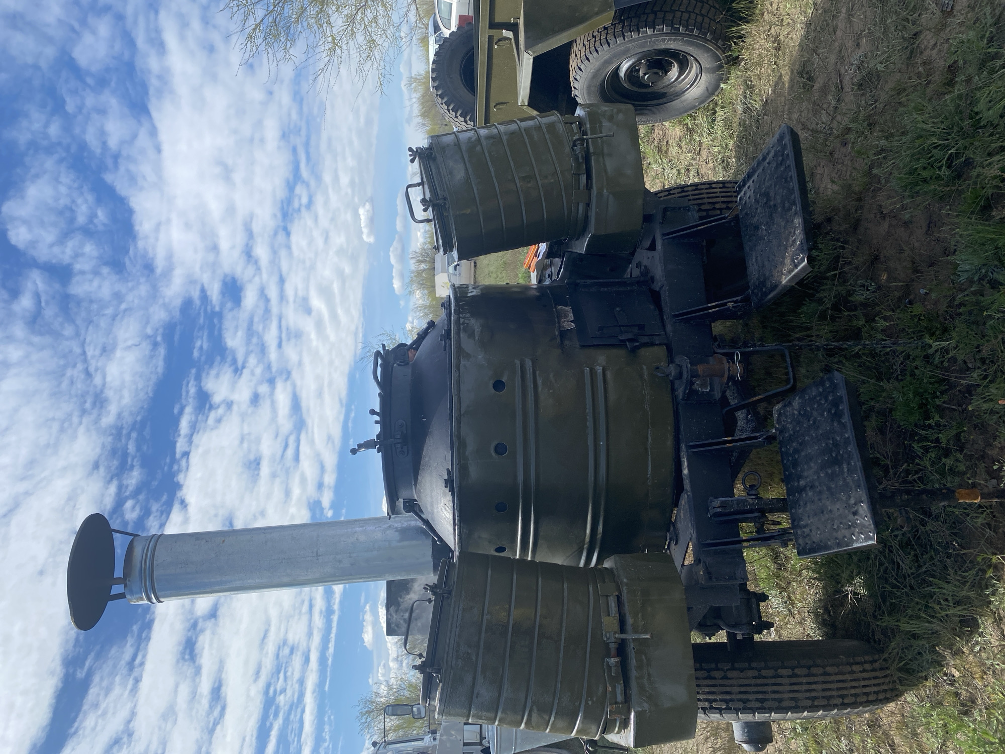The image depicts a military field kitchen set up outdoors, likely on a grassy field near the city of Kumakskoe, Russia. The field kitchen consists of a large, dark green, trailer-mounted structure with several cylindrical containers, potentially cooking vessels, visible on top. A long, silver chimney with a circular cap extends upwards, suggesting it is in use. The structure has what appears to be fold-down metal platforms on the sides. A military vehicle is partially visible in the background. The sky is filled with white, puffy clouds and there is a blue sky behind them.