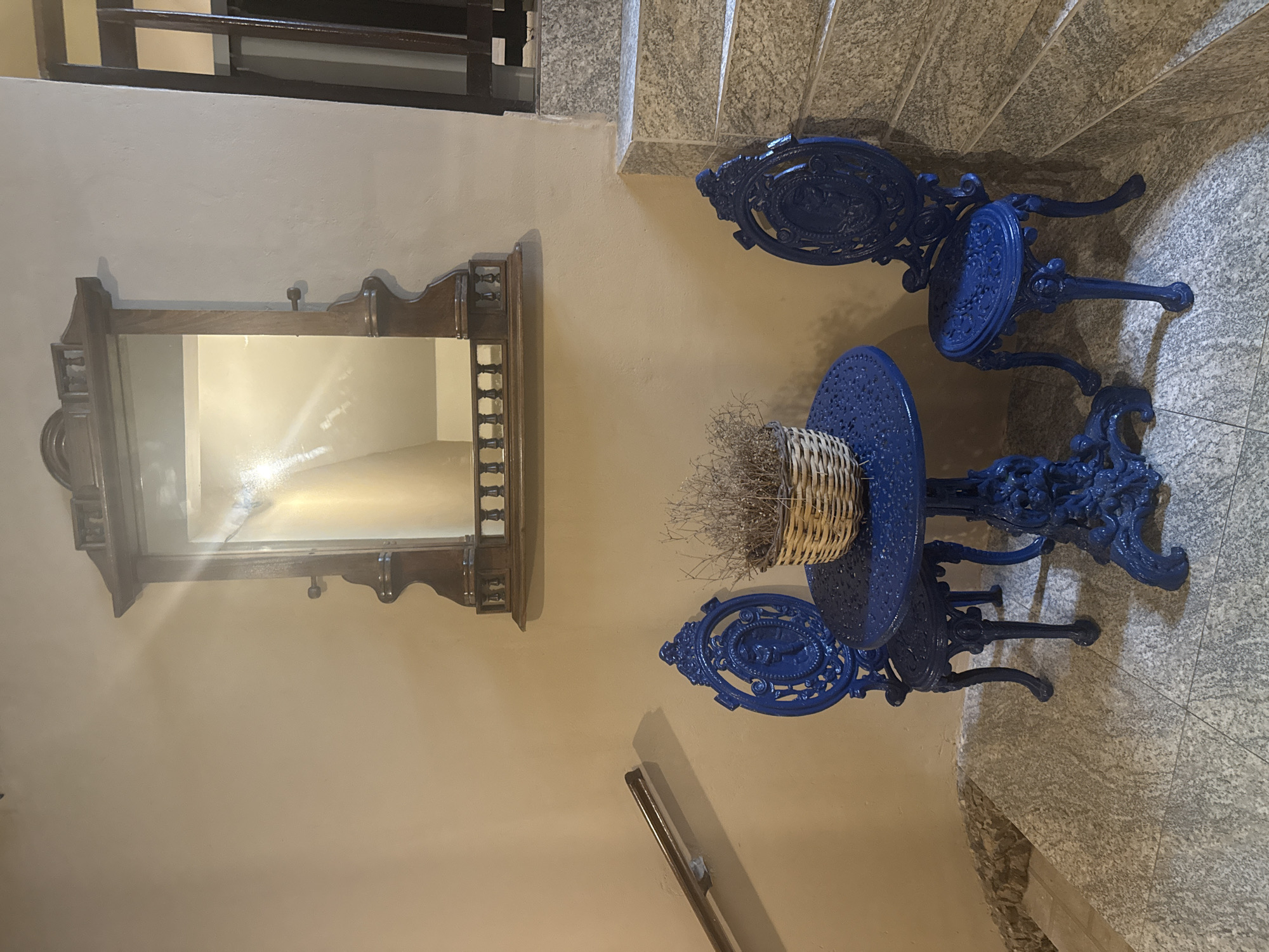 The image depicts an interior scene featuring a small blue wrought-iron table and two matching chairs, arranged on a tiled floor in what appears to be a stairway or hallway. A wicker basket filled with dried foliage sits on the table. Above the table, a decorative wooden mirror with ornate carvings is mounted on a light-colored wall. To the right of the table and chairs, a staircase with granite-like risers is visible. A dark wooden handrail runs along the wall to the left of the scene, above the chairs. The lighting suggests an indoor environment, possibly a residential or commercial building. The specific location in São Pedro Da Aldeia, Brazil, is not ascertainable from the visual information alone. There are no people present in the image, and no discernable actions or interactions are taking place. The overall impression is one of a quaint, somewhat vintage, decorative corner.