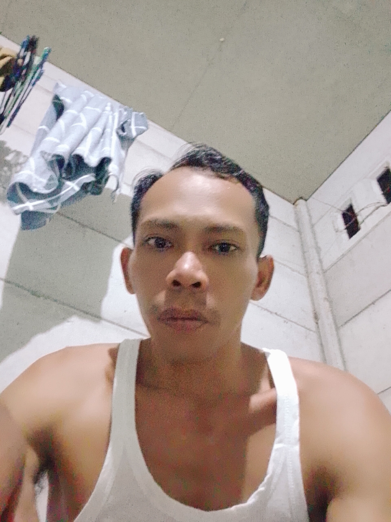 A medium-shot, indoor selfie shows a man in a white tank top. He has dark hair and brown eyes. Behind him, on a wall, is a light gray cloth and a clothes hanger. The wall is light gray with vertical white trim. There are small rectangular vents near the ceiling. The location is the city of Bukit Ketok, Indonesia.