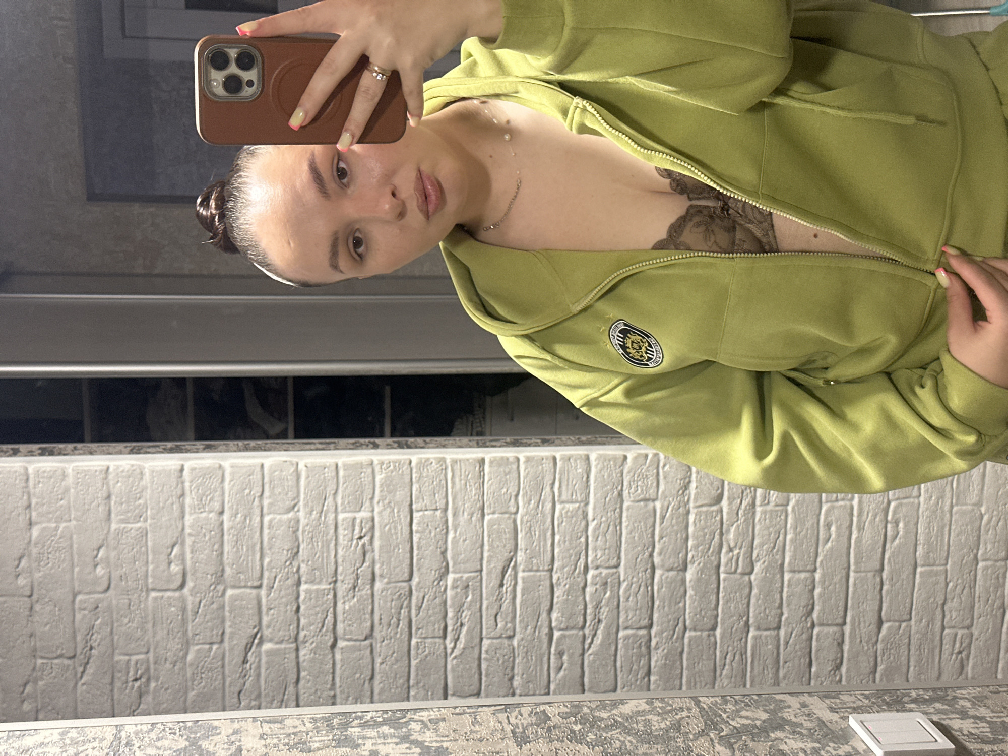 The image shows a person taking a selfie in a bathroom. The person is wearing a green zip-up hoodie, partially unzipped to reveal a lace bra and a delicate necklace. The person is holding a brown smartphone up to take the picture, the phone partially obscures the face. The bathroom has a white faux brick wall and a textured countertop. The location is in Chaikovsky, Russia.