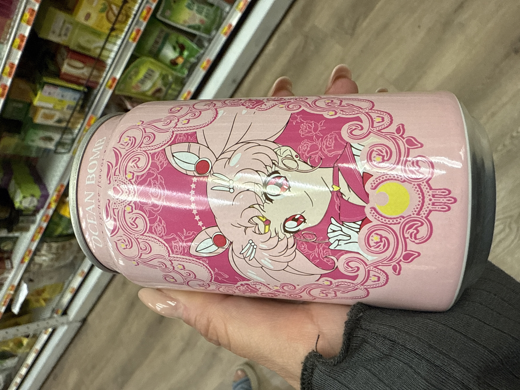 A hand holds a pink can of Ocean Bomb Lychee flavor soda in front of a shelf stocked with groceries. The can features an illustration of Chibi Moon from Sailor Moon, surrounded by an ornate, pink and white design. The background is a shelf of various packaged food items. The setting appears to be a grocery store, possibly located in Davie, United States of America.