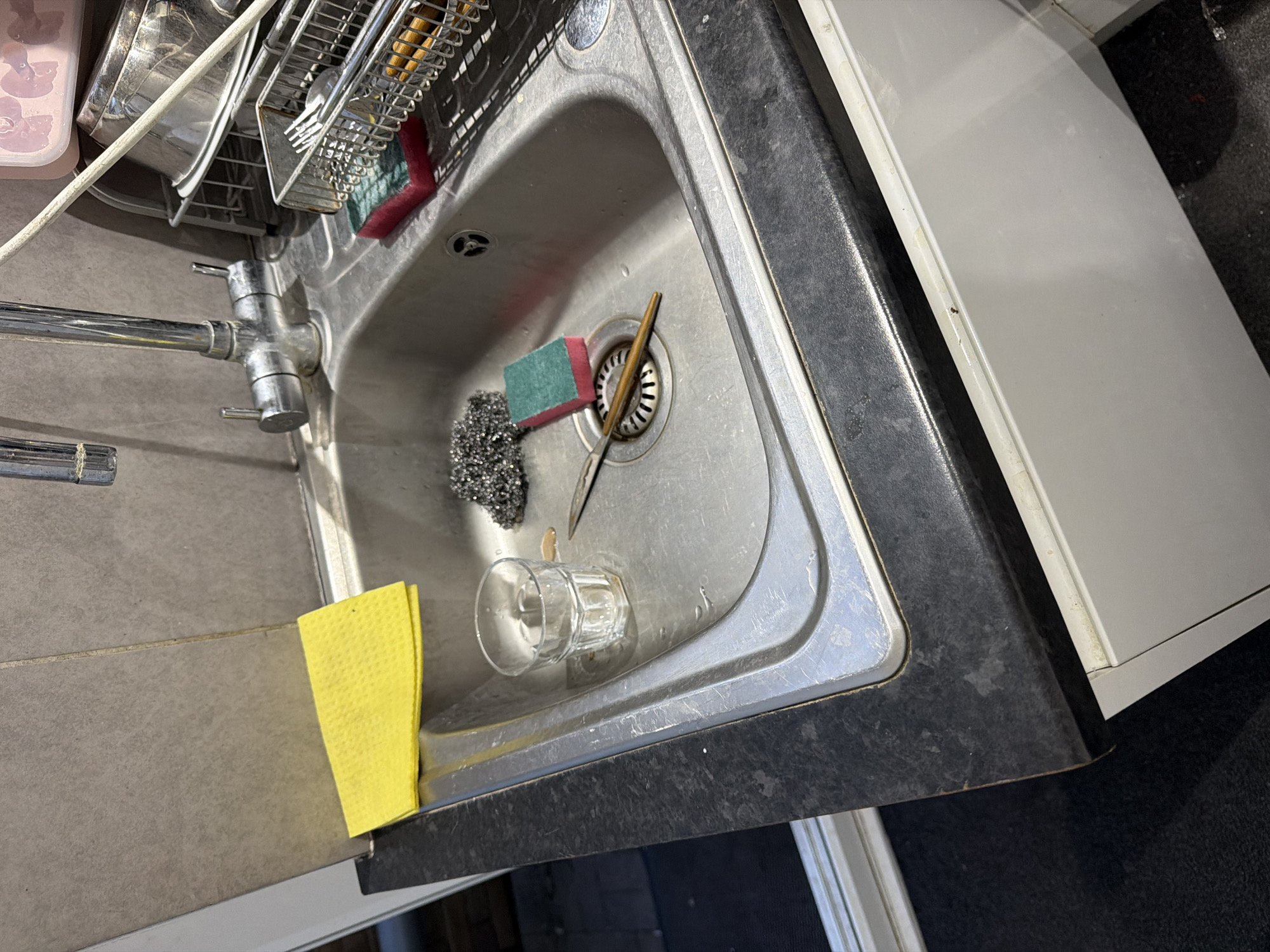The image displays a detailed view of a kitchen sink area. The scene is illuminated by ambient lighting, showing no visible light source.
The central feature is a single-basin, stainless steel sink, presenting visible scratches and water marks indicative of regular use. A chrome dual-handle mixer tap, also showing water spots, is mounted at the rear of the sink. Below the tap, a light gray tiled wall with a vertical grout line extends upwards from a dark speckled countertop.
Within the sink, several items are present: a clear, ridged glass tumbler containing residual liquid stains; a tangled steel wool scrubber; a rectangular, dual-sided sponge with a green abrasive side and a red softer side; and a small knife with a silver blade and a light brown handle, resting across the metallic drain grate.
To the left of the sink, on the dark speckled countertop, a folded yellow absorbent cloth or sponge is positioned. The countertop itself exhibits various water stains and minor wear.
To the right of the sink, a silver-colored metal dish drying rack holds multiple items. It contains a sectioned cutlery holder filled with silver forks and spoons, along with other utensils featuring brown handles. Behind the cutlery, stacked silver-colored metal bowls or pots are partially visible. Resting atop these items is a partially visible plastic container with a pink lid, appearing to contain ice cubes or similar small objects. A white flexible hose or cable extends diagonally from the upper right, passing over the dish rack.
The countertop's edge is visible, meeting white lower kitchen cabinets. To the far left, a dark floor or mat is visible, alongside what appears to be a white door or window frame.
No individuals are present in the image, nor are any actions occurring. The specific geographical location in Maidstone, United Kingdom, cannot be forensically determined from the visual information provided.