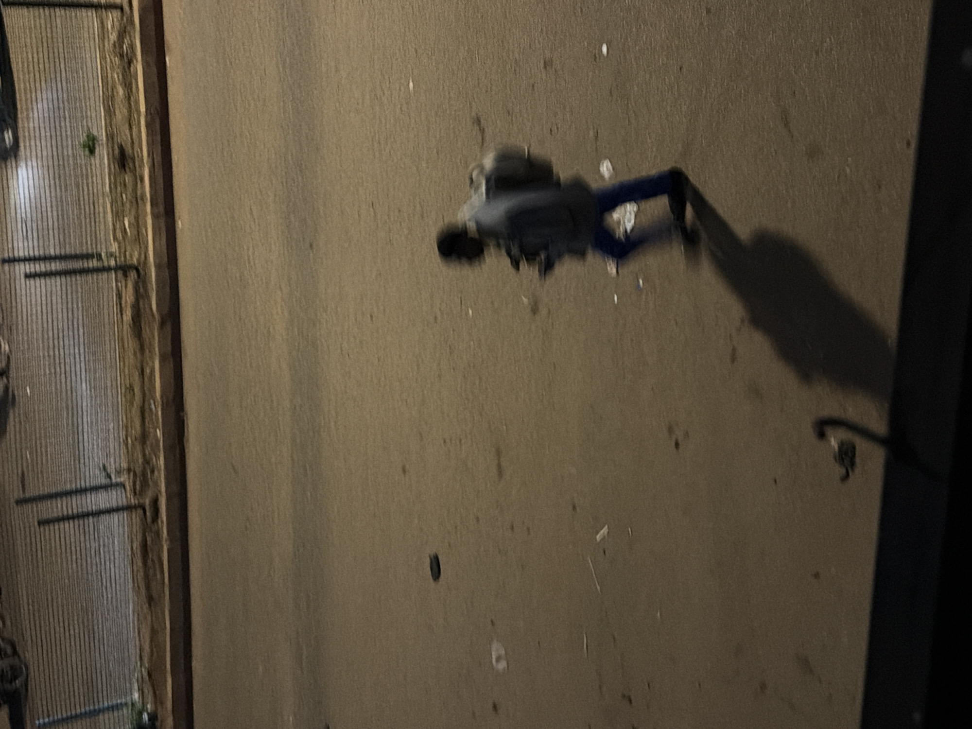The image depicts a person walking on a paved surface, possibly a road or sidewalk, in Bukuru, Nigeria. The individual, with dark skin and wearing a dark jacket and blue pants, is seen from the side and is blurred due to motion. They are carrying a large, light-colored backpack. The ground is sandy and strewn with debris, including white plastic wrappers and other small objects. In the background, a metal fence with horizontal bars runs along a dirt embankment, with some vegetation visible. The scene is dimly lit, suggesting it is nighttime or dusk.