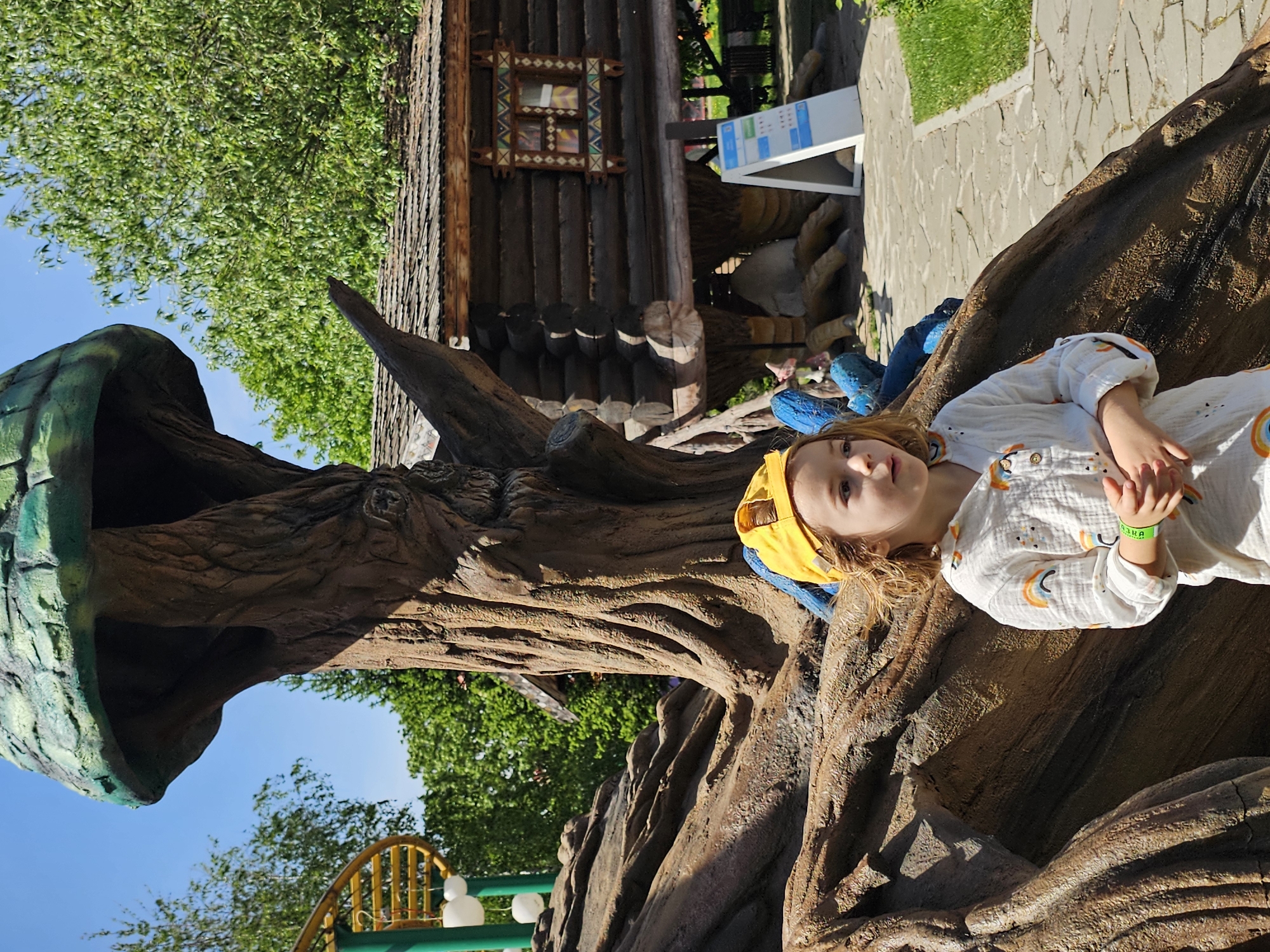 A young child with long blond hair is lying on a tree structure in a park. The child is wearing a white shirt with a rainbow pattern and a yellow hat. The tree structure is made of brown and green concrete and has a large open mouth. In the background, there is a wooden building with ornate window frames and a blue sign. The photo was taken in the city of Khimki, Russia.