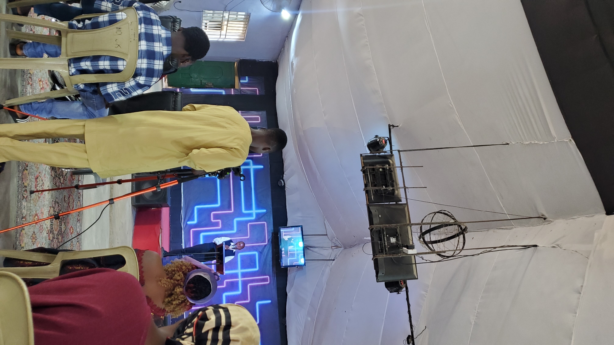 The image depicts an indoor event, likely a service or conference, taking place in Jalingo, Nigeria. The space is covered by a large white tent-like fabric ceiling, supported by metal structures. Overhead, several television monitors are mounted on a metal frame, alongside what appear to be stage lights. A prominent decorative spherical metal object hangs from the ceiling. In the foreground, a man in a yellow kaftan stands facing away from the camera, looking towards a stage. He is positioned near a camera on an orange tripod. To the left, a person wearing a blue and white checkered shirt sits on a plastic chair, facing the stage. Further to the right, a person with curly hair and a purple hat is seated, also facing the stage, and behind them, another person in a burgundy shirt is partially visible. The stage itself features a blue and pink neon light design on the backdrop. A man stands at a podium on the stage, facing the audience and appearing to speak. To the left of the stage, there is a green door and a window with bars. The floor is covered with a patterned rug. The overall atmosphere suggests a gathering with a focus on a speaker, possibly being filmed or broadcast.