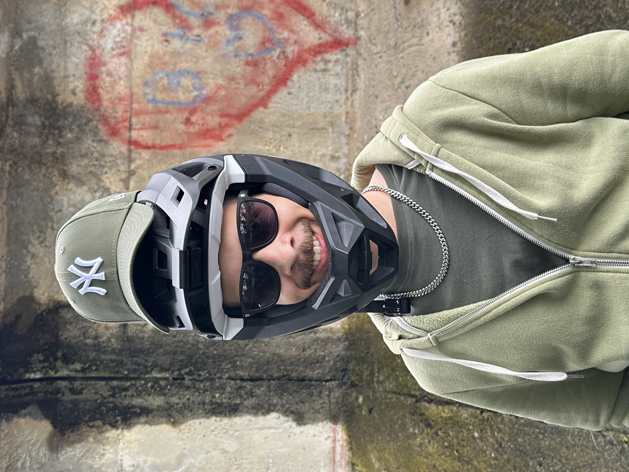 A man stands in front of a concrete wall in what seems to be a public space in Lovech, Bulgaria. The wall is marred with graffiti, including a faded red heart. The man is wearing a green baseball cap with the New York Yankees logo, sunglasses, a black and white mountain bike helmet, a silver chain necklace, an olive green T-shirt, and a light green zip-up hoodie. He is smiling.