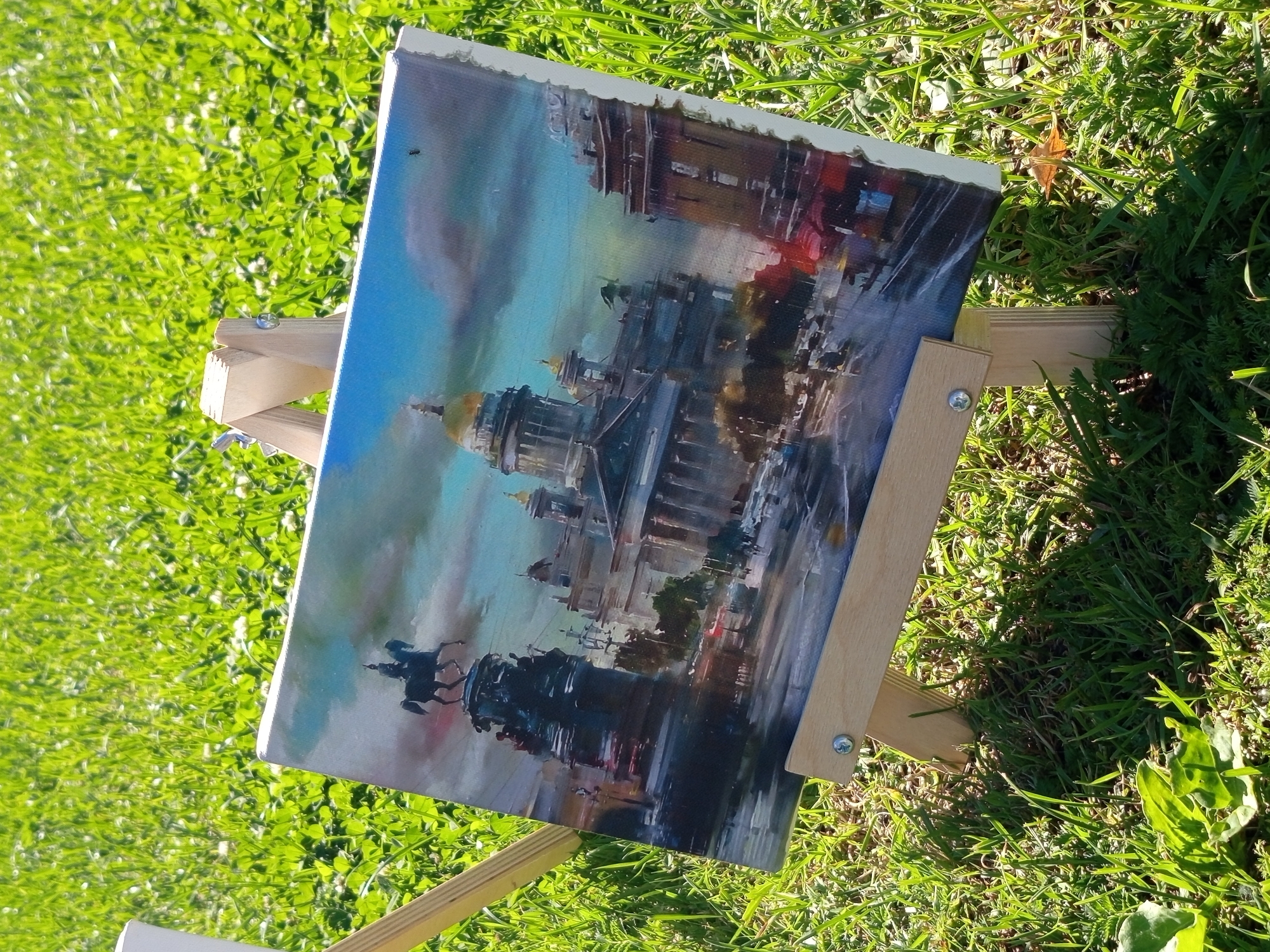 The image shows an oil painting on a wooden easel, placed outdoors on a grassy area. The painting depicts an urban landscape in Saint Petersburg, Russia, specifically the area around Saint Isaac's Cathedral. A statue of an equestrian figure on a tall pedestal is visible on the left side of the painting, believed to be the Bronze Horseman. Saint Isaac's Cathedral, with its prominent golden dome, is depicted in the center. The sky in the painting is a dramatic mix of blues and grays, with hints of sunlight breaking through. The foreground of the painting shows a street scene with a few cars and figures. The easel is made of light-colored wood and is partially obscured by green grass. The background of the photograph is a sunlit expanse of green grass with some clover. There is a small fly visible on the surface of the painting, near the top right edge. The lighting suggests a bright, sunny day.