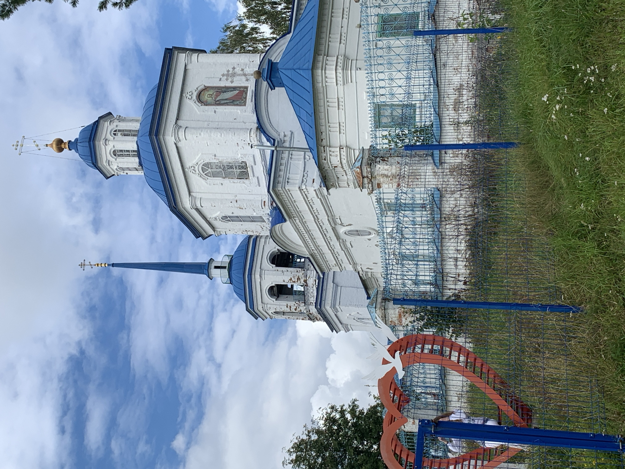 In this image, a white church with a blue roof stands prominently against a cloudy blue sky. The church features multiple domes and a tall spire topped with a cross. A blue metal fence, partially covered in grass, runs across the foreground. To the left, a large red heart-shaped sculpture with white dove figures is visible behind the fence. A person in a white shirt and dark pants can be seen partially within the heart sculpture. The environment appears to be overgrown with grass and some trees visible in the background. The scene is captured from a low angle, looking up towards the church.