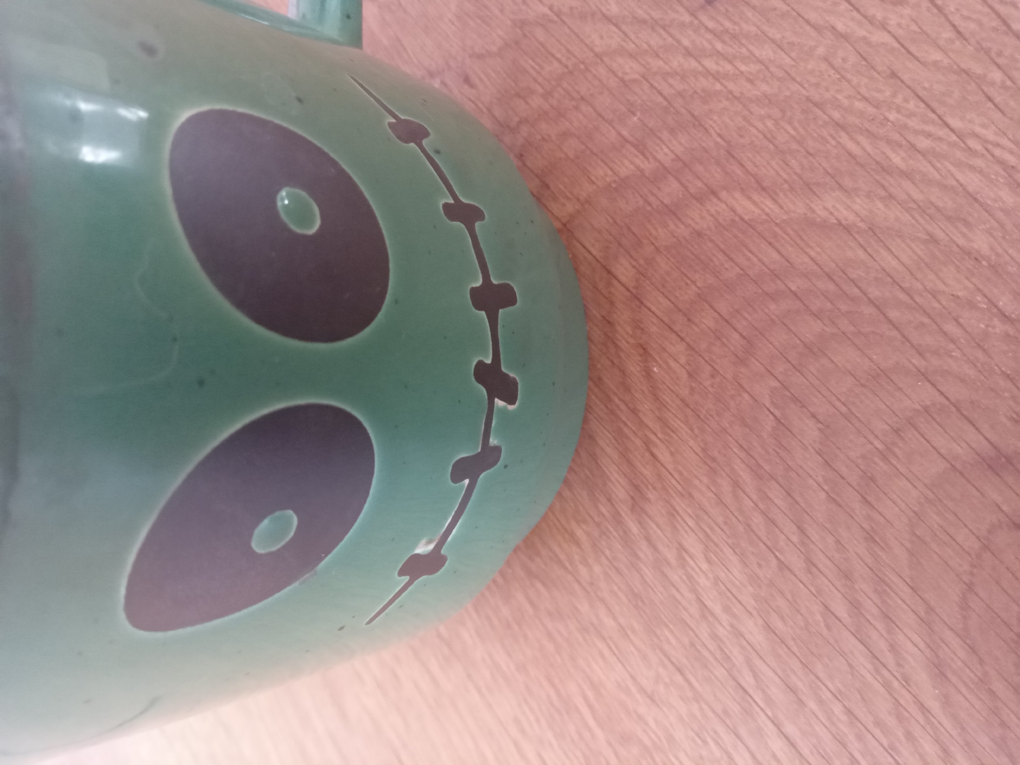 A close-up, eye-level shot shows a green ceramic mug with a Halloween-themed design on a light brown wooden surface. The mug features two large, dark oval eyes, each with a smaller white oval inside, giving it a cartoonish, spooky appearance. A stitched mouth, also in dark, completes the face. The mug has a handle visible on the right side. The wooden surface has a visible grain pattern. The image does not contain any information about the city of Terebovlia, Ukraine.