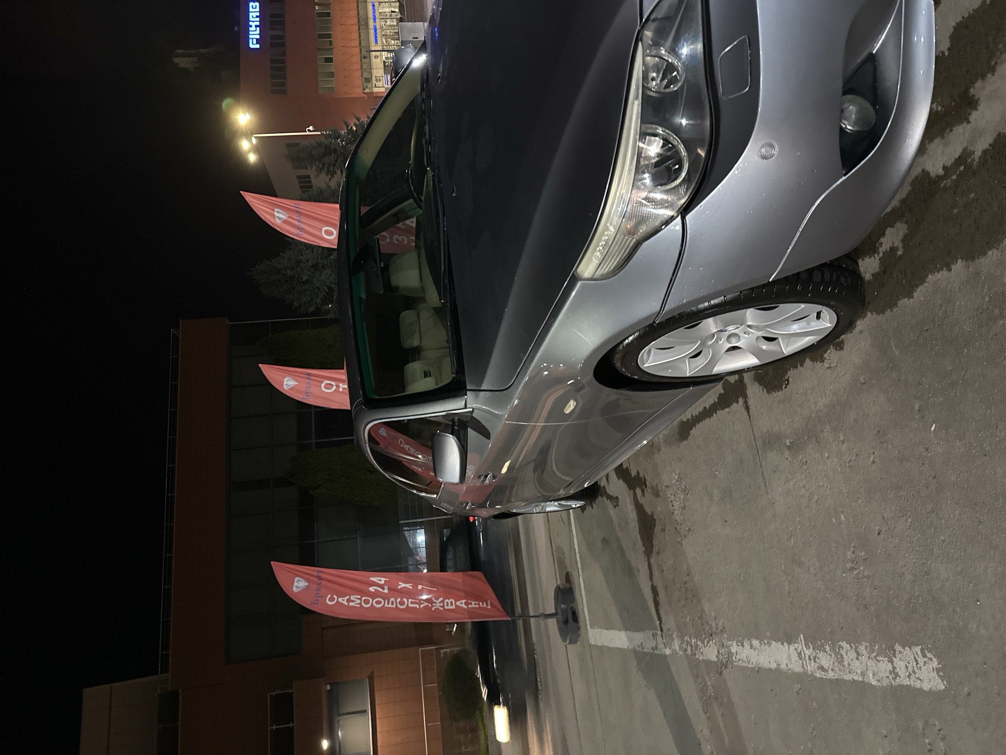 A silver car is parked on a paved surface in front of a building with signage visible, possibly in the city of Plovdiv, Bulgaria. Several red flags with writing in Cyrillic stand near the car. The scene appears to be at night or in low light conditions, with street lights and building lights illuminating the area. The car is parked in a designated parking space.