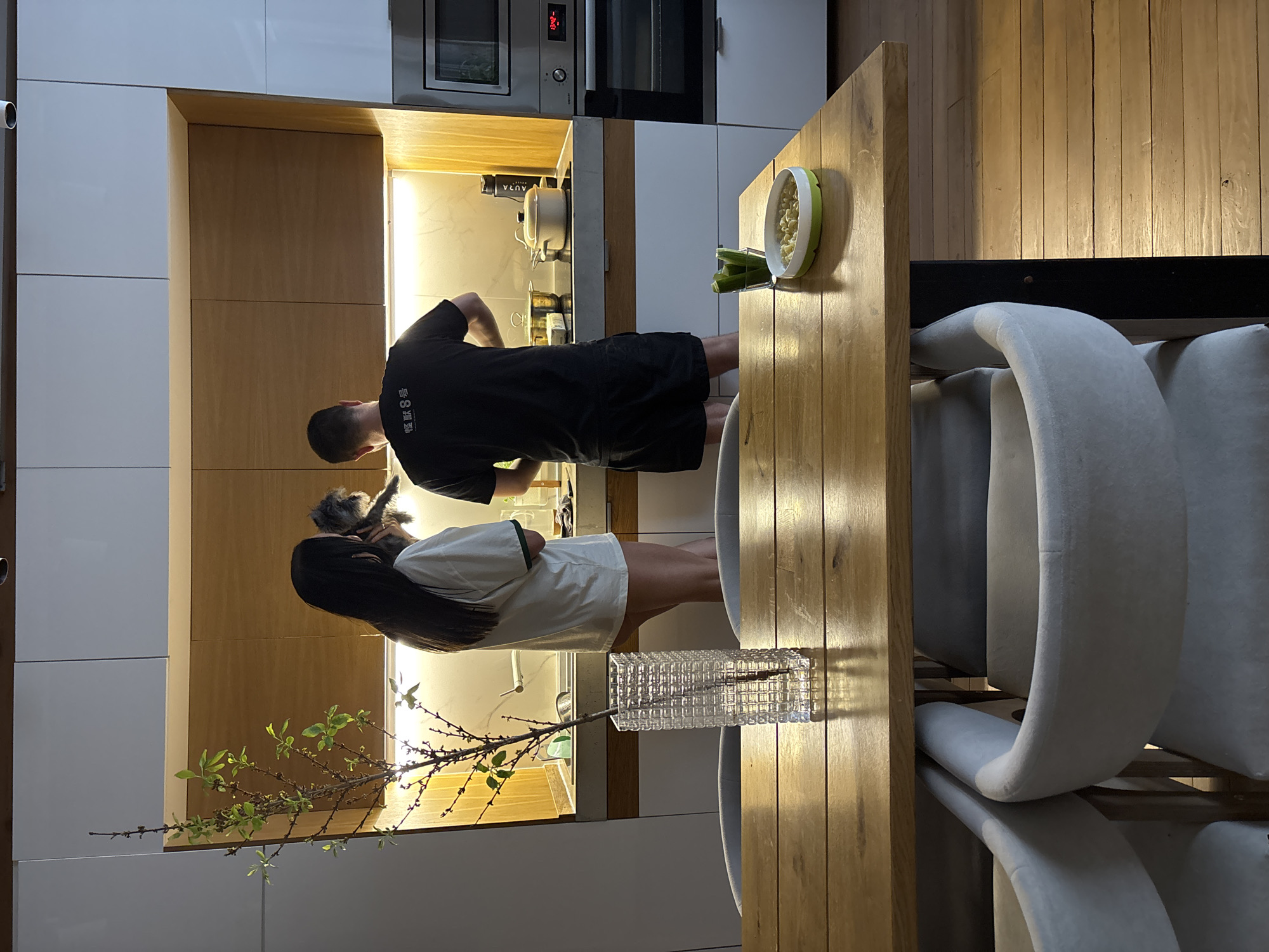 A man and a woman are in a kitchen. The man is wearing a black t-shirt and black shorts and is standing at the counter. The woman is wearing a white shirt and shorts and is holding a dog. There is a wooden table with two chairs. On the table, there is a bowl of food and a vase with flowers. The kitchen has white and wood cabinets and stainless steel appliances. The scene appears to be a domestic setting. The location in the city of Chaikovsky, Russia is not deducible from the image.