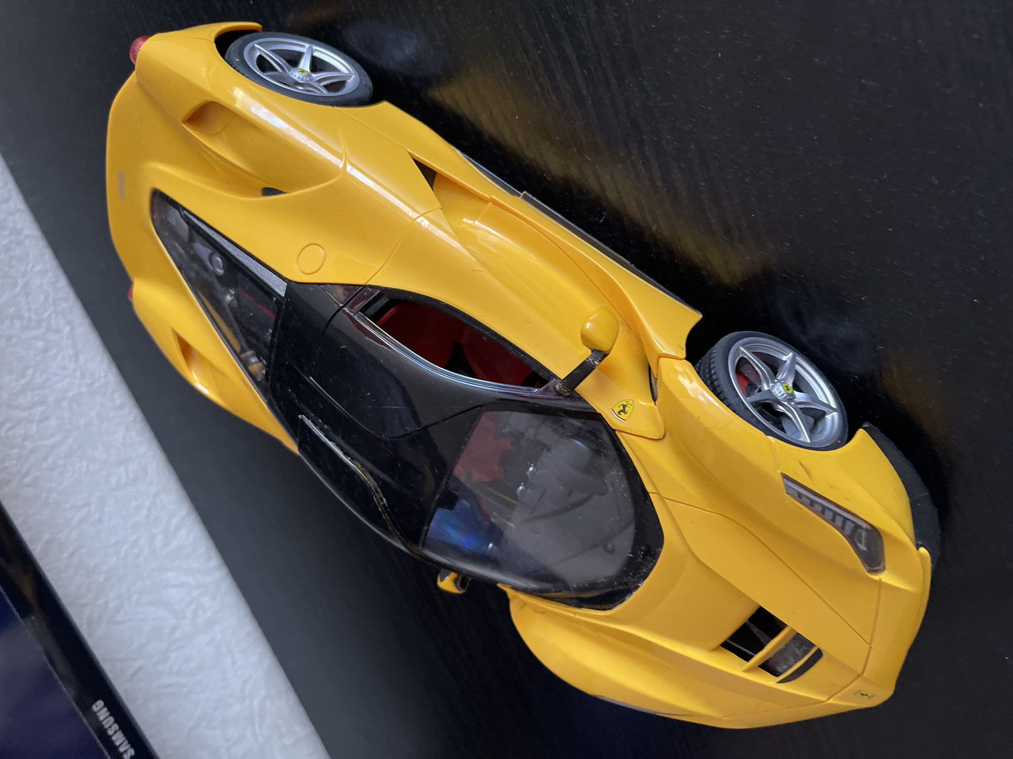 The image features a detailed, bright yellow model car, strongly resembling a modern Ferrari sports car, displayed on a dark, reflective surface. The car is positioned diagonally across the frame, with its front angled towards the bottom right and its rear towards the top left.
The model car boasts intricate details, including multi-spoke silver wheels with visible red brake calipers. Its windows and roof are a dark, tinted black, allowing a glimpse of the red interior. A small, iconic Ferrari "prancing horse" emblem is visible on the side of the car, near the base of the windshield. The car's design includes sleek headlights with horizontal elements, side mirrors, and prominent grilles on the front bumper. The yellow body shows minor surface dust, suggesting it is a cherished display item.
The background consists primarily of the dark, semi-glossy surface the car rests upon. To the far left, a strip of a white, textured surface is visible, alongside a dark object that bears the white text "SAMSUNG" in capital letters, likely a book, tablet, or another electronic device.
The scene depicts a stationary object in an indoor setting, likely on a table or desk. There is no activity taking place, and no people are present. The lighting suggests an indoor environment, and the time of day or external weather conditions are not discernible from the image. The visual content does not provide specific cues for its location in Moscow, Russia.