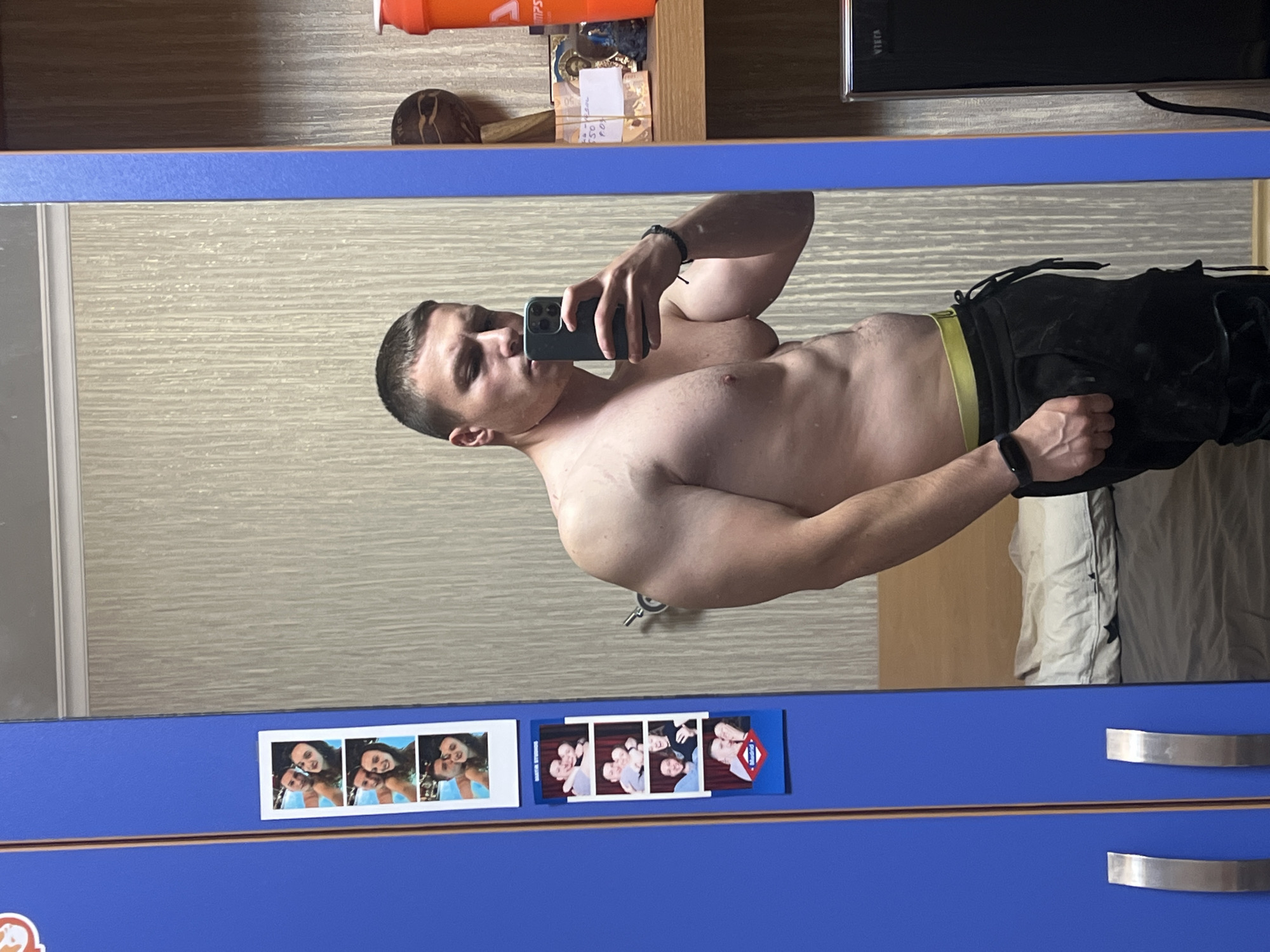 A shirtless man is taking a selfie in a mirror. He is wearing dark athletic pants with a visible yellow waistband and a black wristband. The mirror is attached to a blue dresser with silver handles. There are two strips of photo booth pictures stuck to the dresser below the mirror. The background shows a shelf with various objects including a protein shaker, a small decorative item, and some papers. The wallpaper behind the mirror is neutral-toned with a textured pattern. The location appears to be indoors, possibly a bedroom. The city of Burgas, Bulgaria, is not deducible from the image.