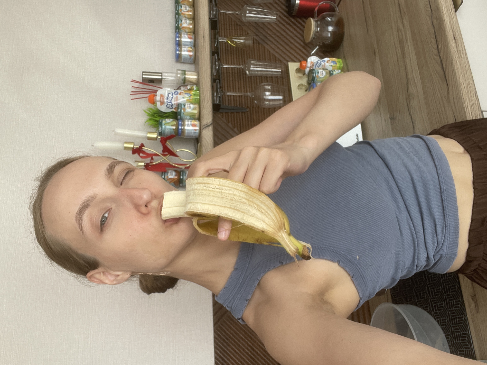 A woman with fair skin and light brown hair, pulled back in a low bun, is eating a banana. She is wearing a light blue ribbed tank top and brown bottoms. She is winking at the camera. Behind her, a wall is visible, with a set of shelves containing bottles and jars of food. The location is Подшивалово, Russia.