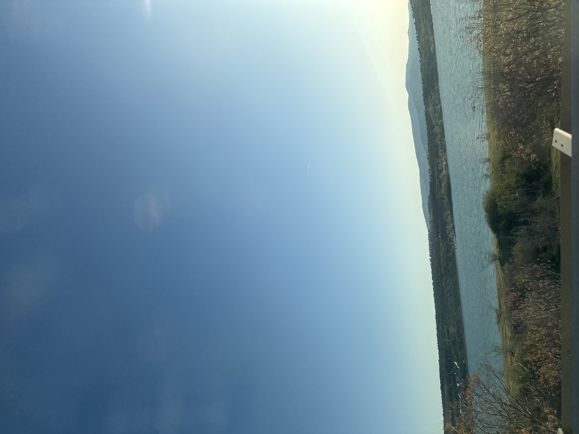 The image depicts a view of a body of water, likely a lake or river, flanked by land with vegetation. Mountains can be seen in the distance along the shoreline. A portion of the sky is visible, transitioning from a darker blue to a lighter hue near the horizon. Some trees and shrubs are in the foreground, near what appears to be a barrier or railing. The orientation suggests the image was taken at an angle. While it's a landscape view, specific details that would pinpoint the location within Tbilisi, Georgia, are absent.