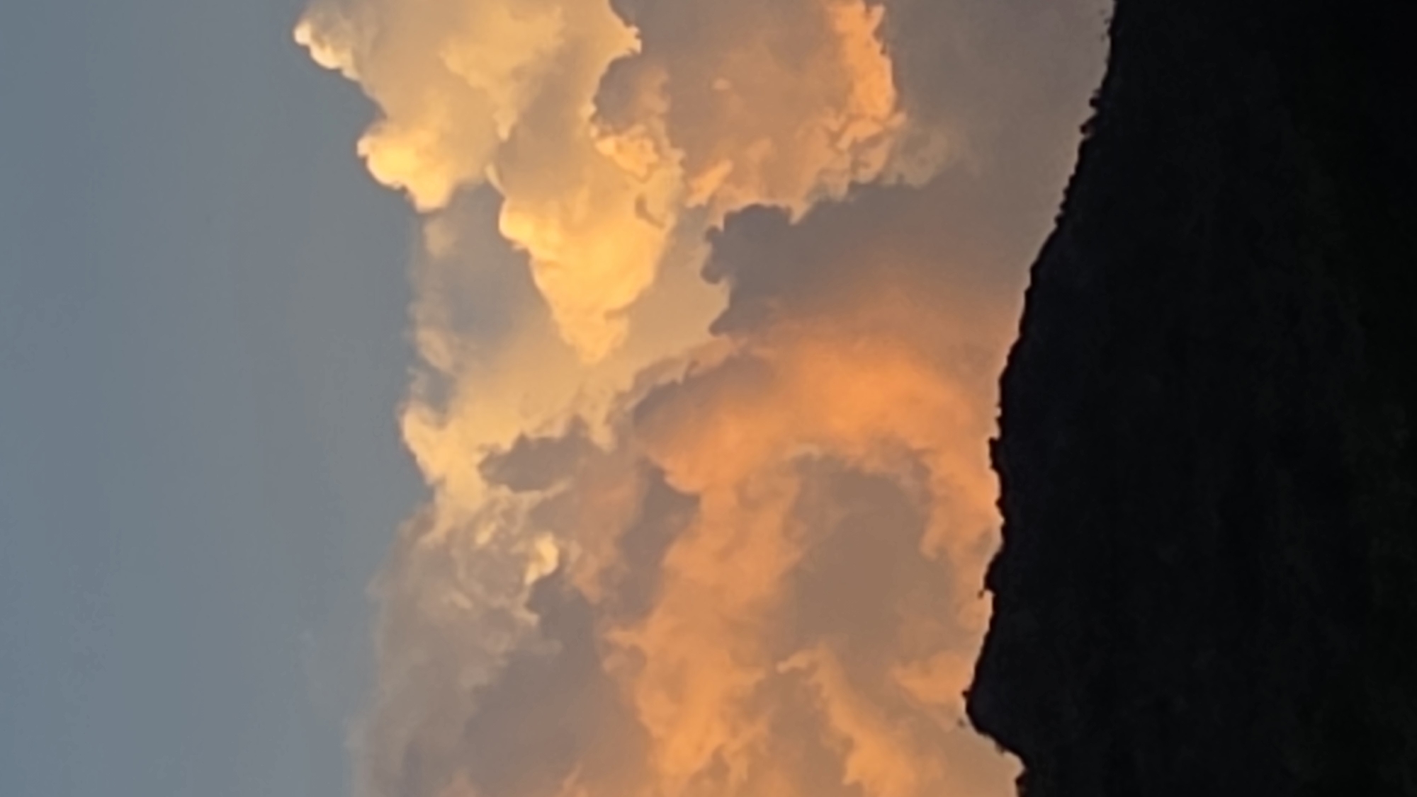 The image shows a landscape at sunset or sunrise, potentially in the city of Bac Nha Trang Ward, Vietnam. A large, dark, possibly mountainous landmass occupies the right side of the frame. To the left of this landmass, the sky is filled with vibrant, golden-orange clouds, suggesting either a sunrise or sunset. The remaining sky is a lighter blue-grey color. The composition focuses on the interplay between the dark land and the colorful clouds.