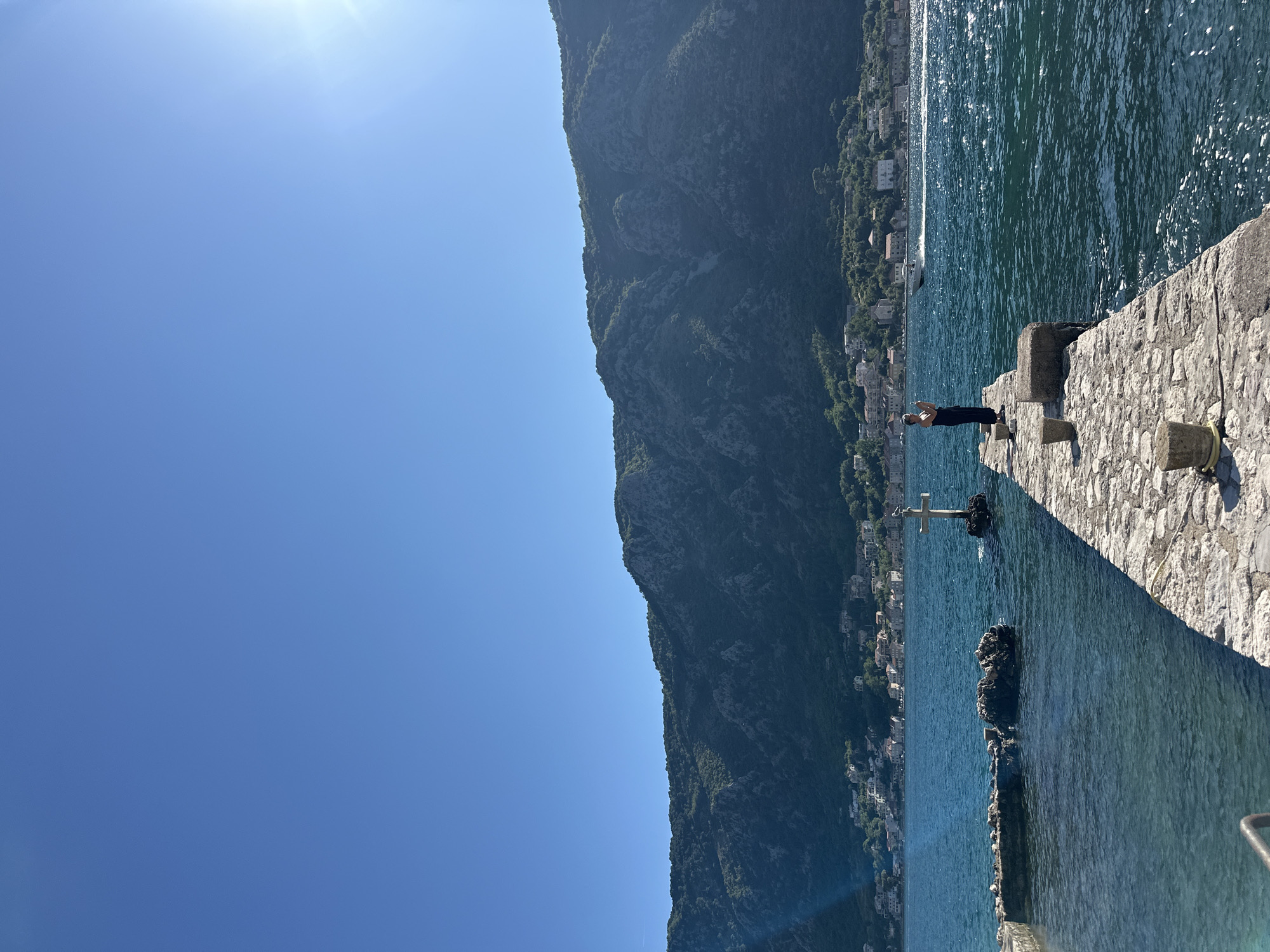 A lone person stands on a stone pier extending into a bright blue sea. The person is wearing dark clothing. Behind the pier, a town with white buildings is nestled at the base of lush green mountains. The sun is high in the sky, casting a bright glare on the water. A small boat is visible in the distance. A cross-shaped structure stands in the water near the pier. The scene appears to be a scenic coastal area, likely the town of Dobrota in Montenegro, known for its beautiful bay and mountainous landscape.