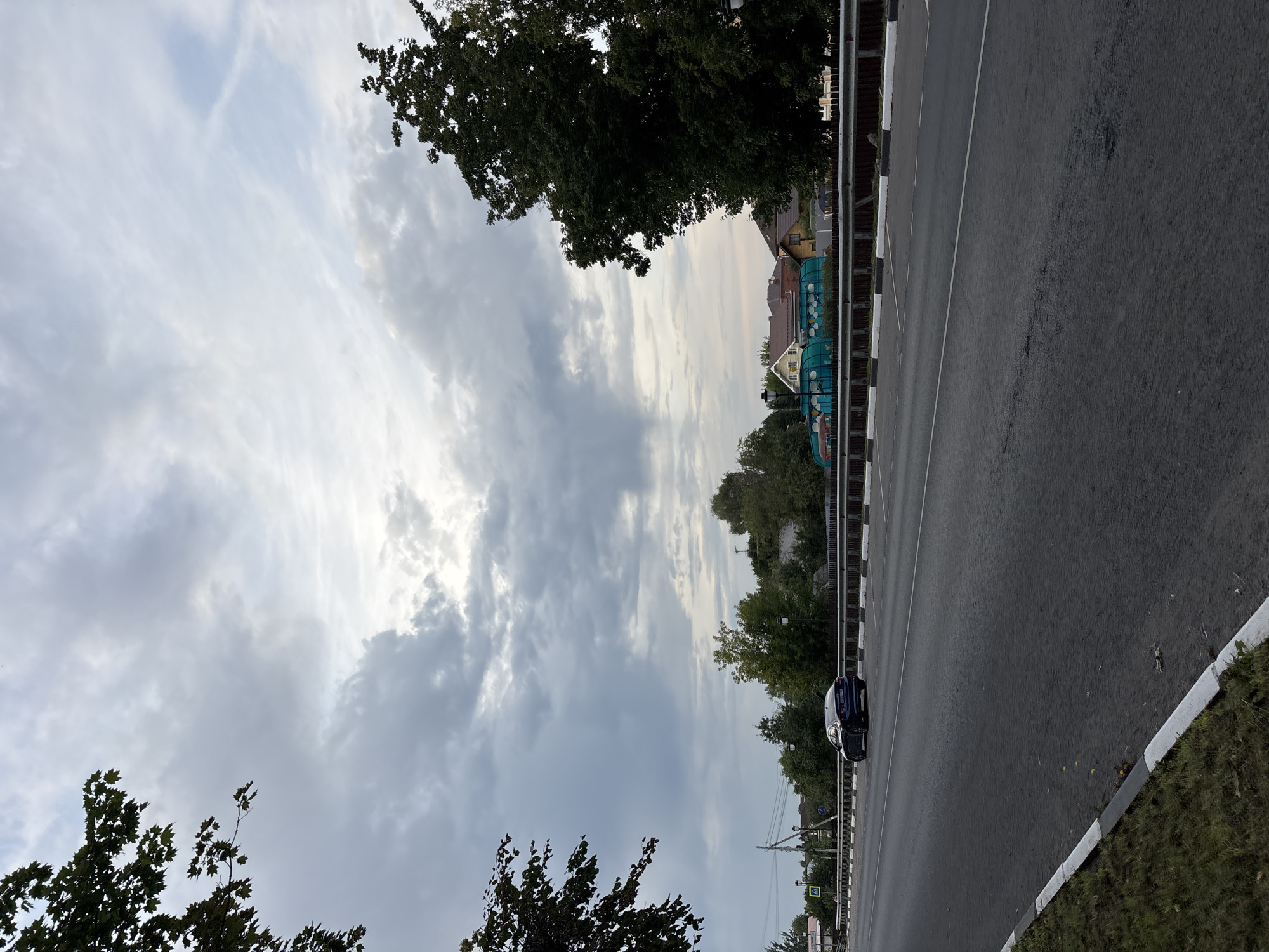 This is a horizontal, eye-level, outdoor, mid-range shot of a road in Klin, Russia. The sky is filled with gray, overcast clouds, with patches of lighter sky visible. The foreground consists of a dark asphalt road with a white and black painted curb and grass on the left side. A dark blue sedan is driving on the right lane of the road. In the middle ground, there is a metal guardrail separating the road from a grassy area with trees. Behind the guardrail, there are several houses with red and brown roofs, and some green structures that appear to be children's play areas or enclosures. Tall trees with dark green foliage frame the top left and right sides of the image.