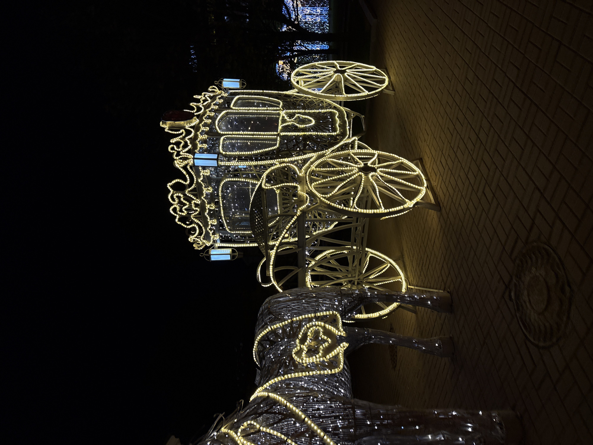 The image captures an illuminated decorative carriage and horse in Minsk, Belarus. The carriage, outlined with bright lights, has four wheels and is intricately designed with scrolling details. A horse figure made of similar lights is hitched to the carriage. The scene is set at night on a brick-paved area, and the background is dark.