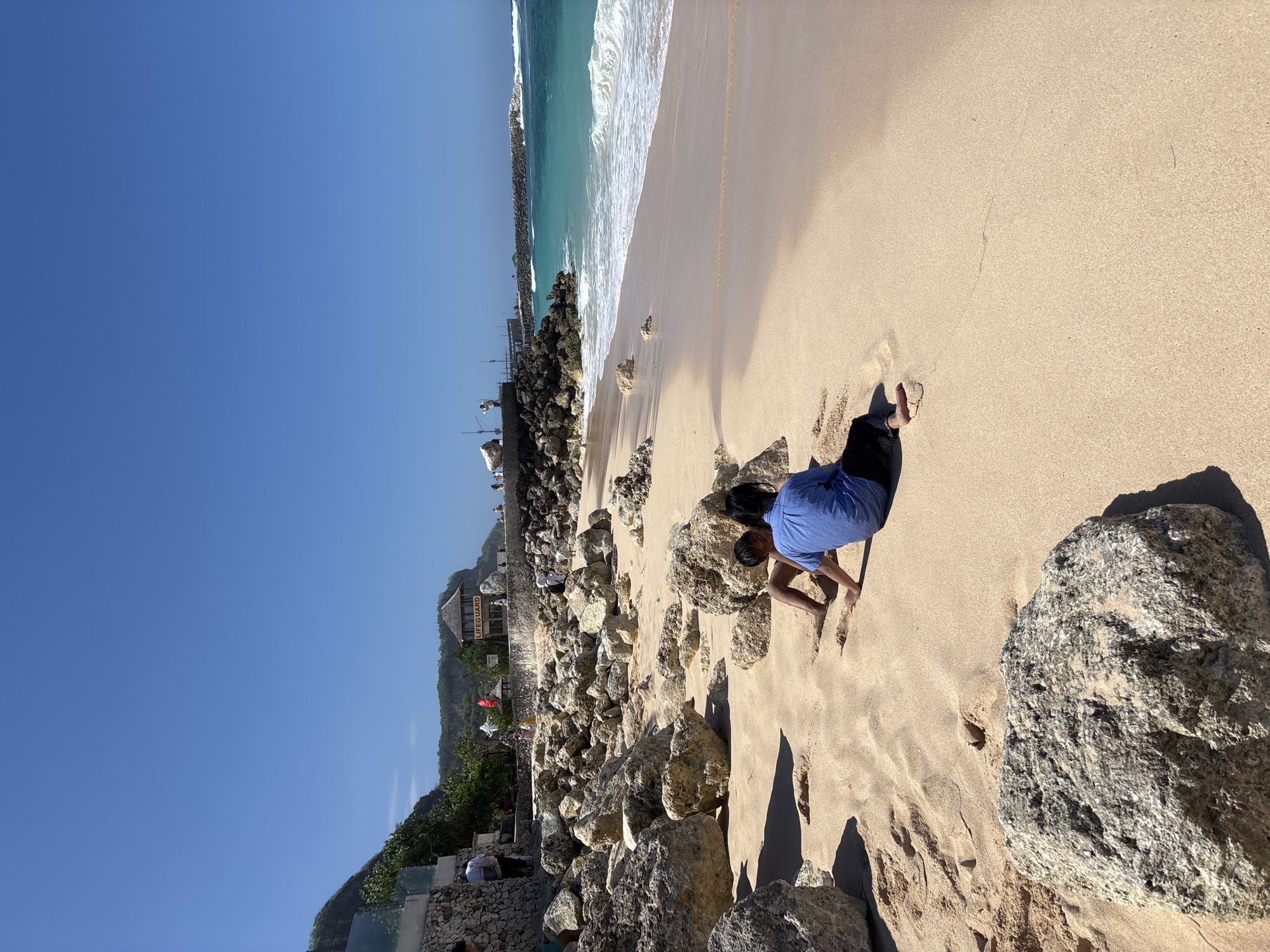 The image captures a sunny day at a beach in Kuta Selatan, Indonesia, likely in the vicinity of the cliffside resorts and restaurants overlooking the ocean. The foreground is dominated by fine, light-colored sand with scattered large rocks. Two individuals, likely children, are seated in the sand facing away from the camera, one wearing a blue shirt and black pants, the other is mostly obscured. Their interaction appears casual, possibly playing or resting.
In the middle ground, the sandy beach transitions to the turquoise ocean with gentle waves lapping the shore. Beyond the beach, a rocky embankment is visible, topped with a low wall. Further back, a lifeguard tower with a red roof and the word "LIFEGUARD" in white lettering is prominent, suggesting an area designated for swimming and safety. Several other people are scattered along the embankment and on the beach, some walking, others observing. The background features a lush green cliffside rising behind the man-made structures, with more distant hills under a clear blue sky. The overall scene depicts a popular tourist destination with a relaxed beach atmosphere.