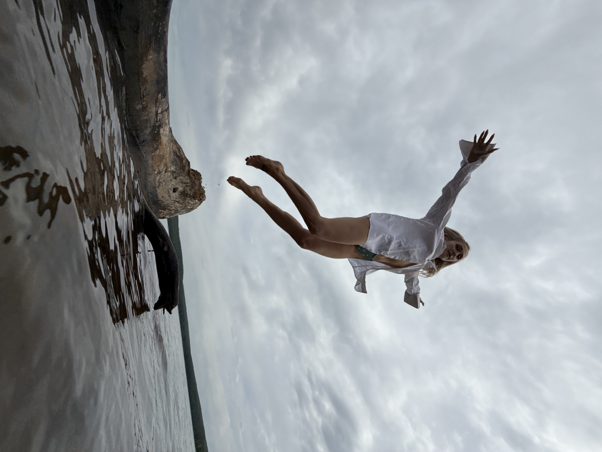 A woman in a white shirt leaps from a log over the water, likely a lake or river. Her arms are outstretched, and her legs are extended behind her. The sky is overcast with cloudy skies. The left side of the image shows the water and the log, with its reflection visible in the water. Based on the general aesthetic and the possibility of being somewhere in Russia, it's impossible to confirm if this image was taken in Chaikovsky, Russia, based only on this image. Therefore, information regarding the location is not provided.