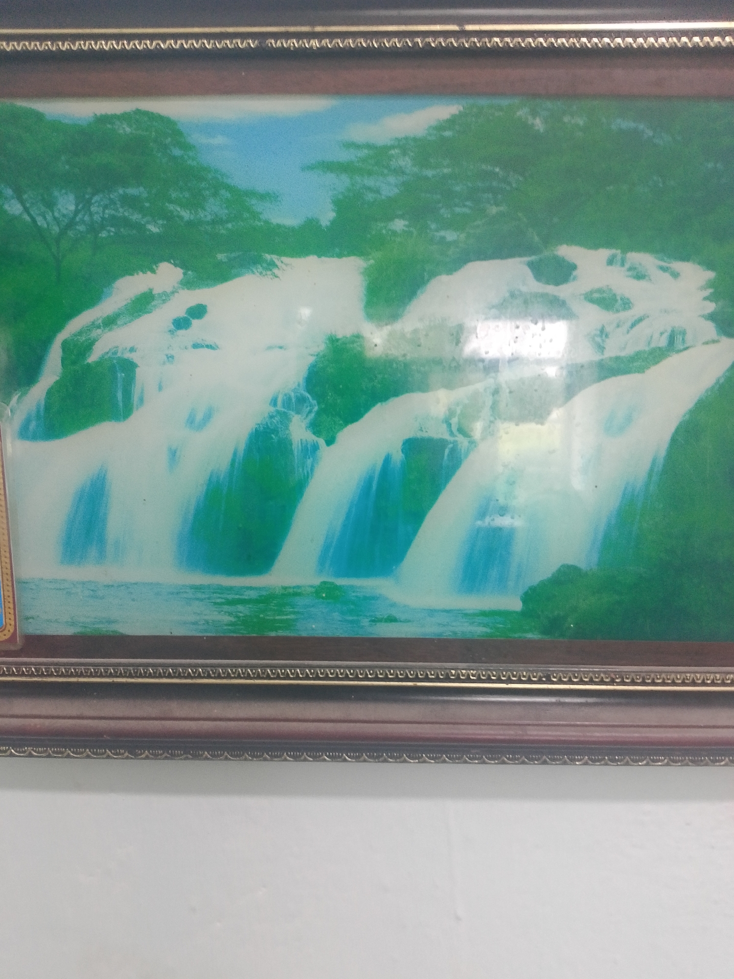 A framed painting of a cascading waterfall is displayed on a white wall. The painting depicts a lush green landscape with trees and a bright blue sky. The waterfall itself is rendered in shades of white and blue, with water flowing over rocks into a calm body of water at the bottom. The frame of the painting is dark brown with decorative metallic trim. The image is slightly blurry and there are some reflections on the surface of the painting. The location in the city of Asino, Russia, is not visible in the image.