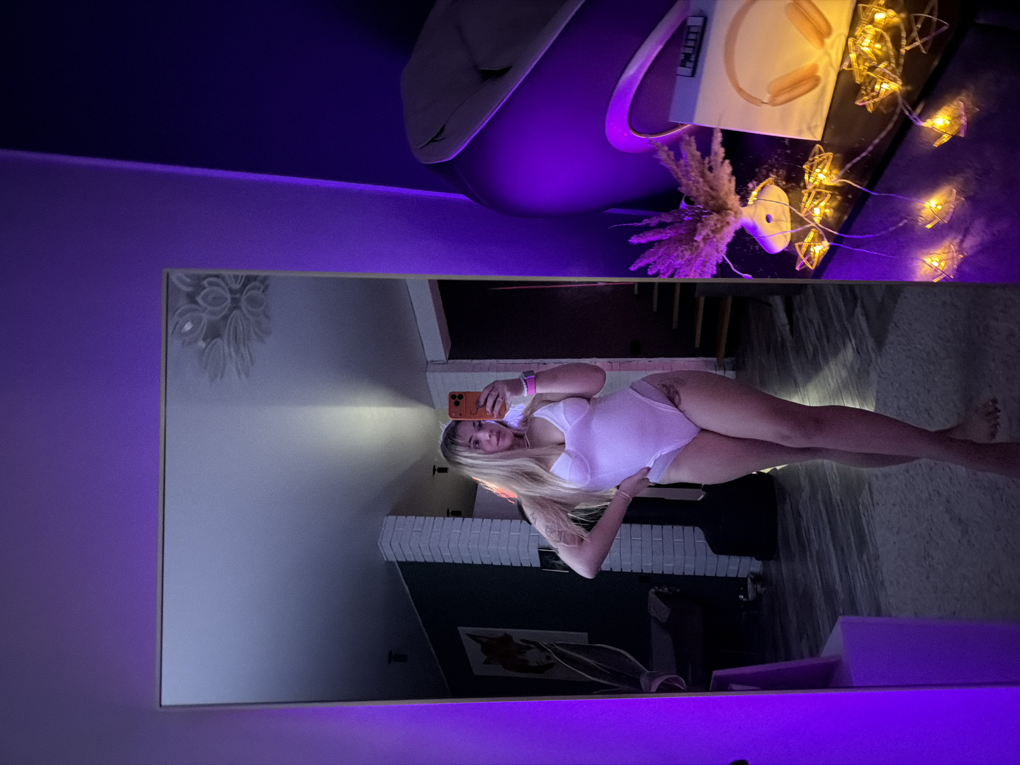 A woman is visible in the reflection of a rectangular mirror mounted on a wall in a room. She is holding a smartphone in her right hand, seemingly taking a selfie. She is wearing a white bodysuit and has blonde hair. The reflection behind her shows a living room with a fireplace, stairs, artwork on the wall, and furniture. The mirror is placed on a wall that is illuminated with purple light. On a surface above the mirror, there is a chair, headphones, decorative lights, and a plant. The location is identified as Большая Шильна, Russia.