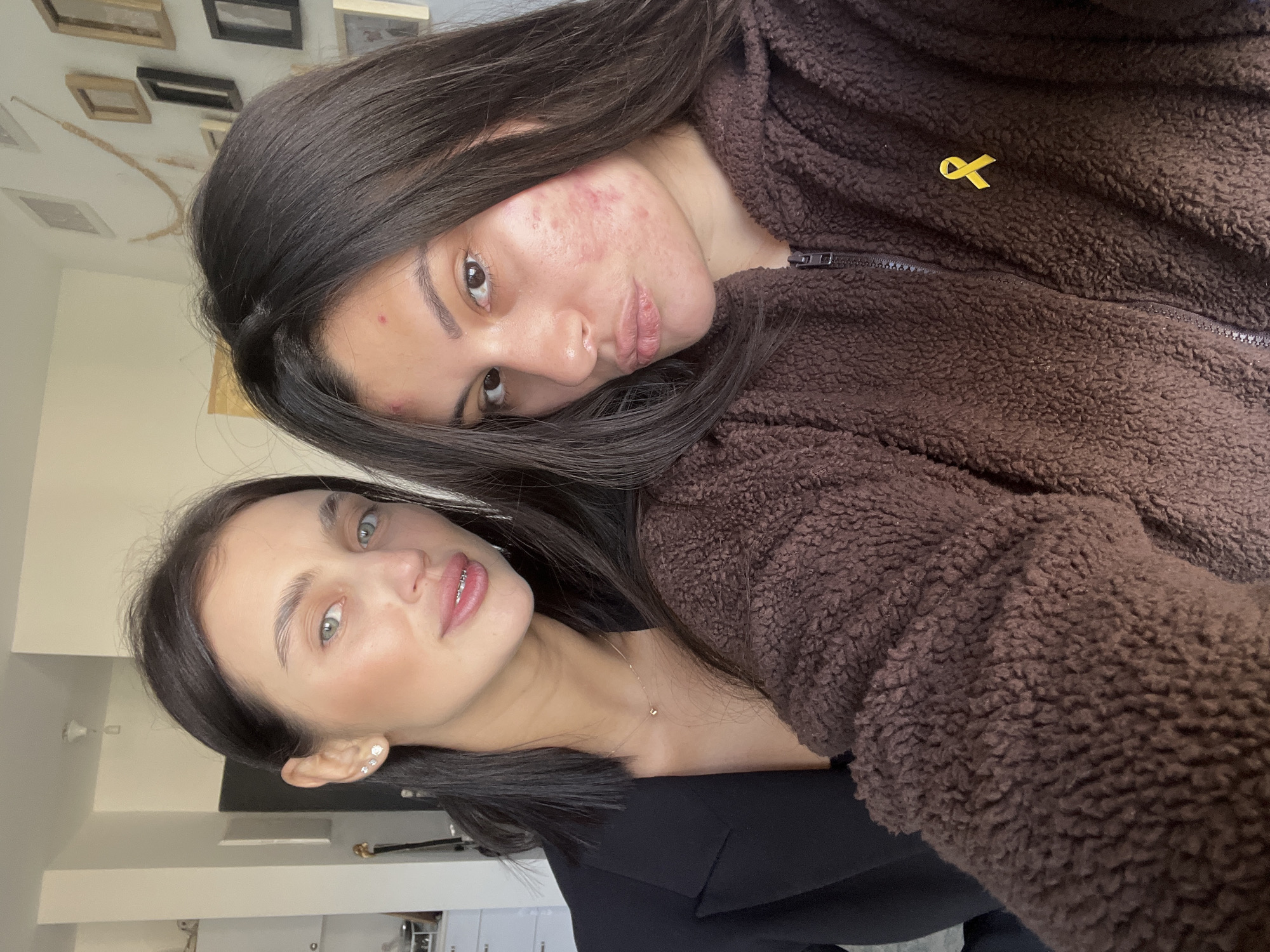 This close-up selfie shows two young women positioned closely together in an indoor setting. The image is rotated 90 degrees counter-clockwise from a typical upright orientation.
In the foreground, a woman with dark hair and light eyes looks towards the camera with a slight pout. She has fair skin, visible dental braces on her lower teeth, and multiple small stud earrings. A delicate gold-colored necklace with a tiny pendant adorns her neck. She is wearing a dark, likely black, top.
Behind her, another woman with dark hair and eyes also looks towards the camera. Her left cheek has noticeable redness and acne. She is wearing a brown, fuzzy jacket, possibly fleece or sherpa, which features a zipper down the front. Pinned to her jacket is a small yellow ribbon.
The background consists of a light-colored wall decorated with several framed items. The lighting suggests an indoor environment. The women appear relaxed and are posing for the photo. The location context for this image is Kiryat Bialik, Israel. No visible text is present in the image.