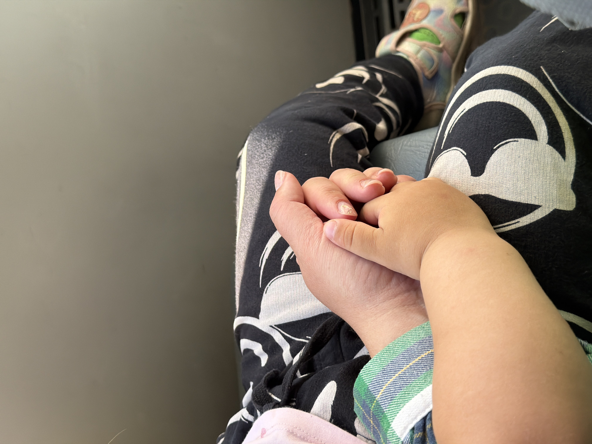 The image shows two people holding hands in what appears to be a public setting, possibly a bus or train. The person on the right is wearing a green and blue plaid shirt with their arm resting on the person on the left. The person on the left is wearing black pants with white graphics and colorful sandals. The background is a plain gray wall. There is no information in the image or provided to confirm the location of the city of Chaikovsky, Russia.