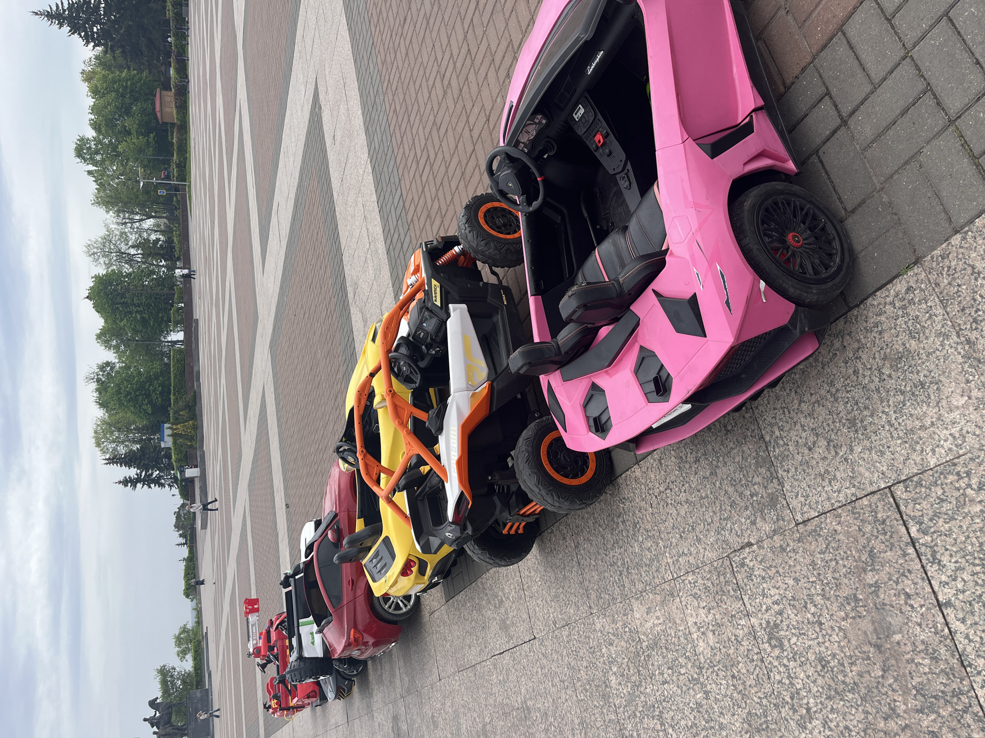 A row of toy vehicles sits parked on a paved plaza in Ulyanovsk, Russia. The plaza's surface is composed of a geometric pattern of interlocking tan, brown, and white pavers. The vehicles, arranged in a line that recedes into the distance, range in color and style, including red, yellow, white, orange, and pink. The vehicles nearest to the camera appear to be children's ride-on toy cars. A pink Lamborghini toy car is closest to the camera.
In the background, trees and shrubs line the edge of the plaza. Figures of people can be seen standing and walking in the distance along the edge of the plaza. The sky is overcast. The overall scene is an urban public space with children's toy vehicles as a central element.