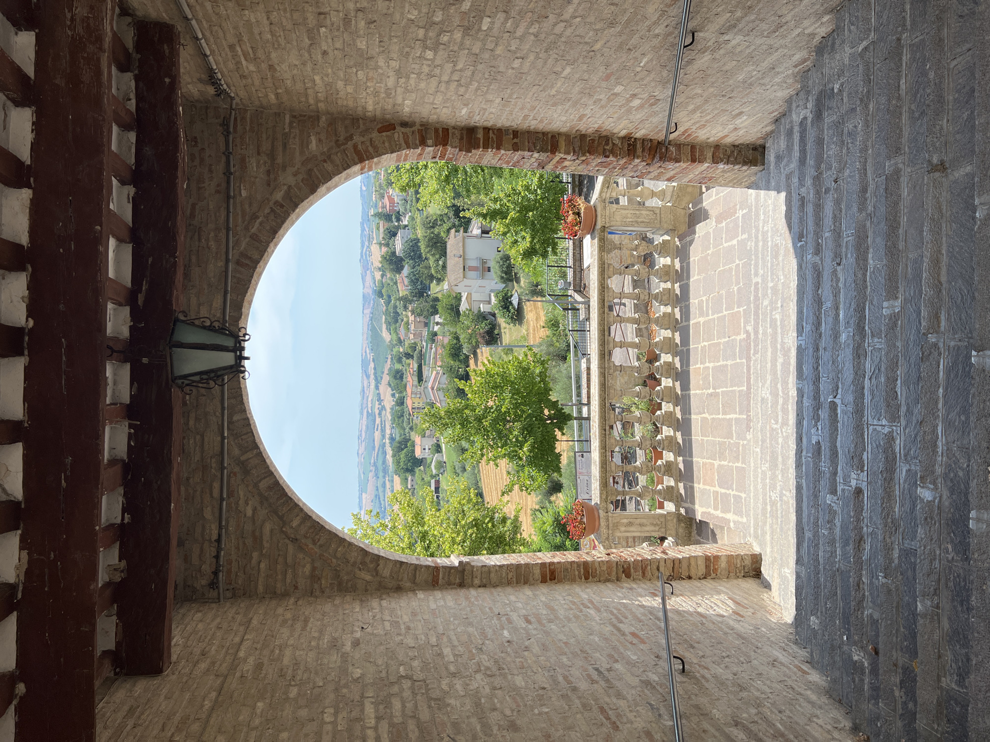 The image shows a view through an arched brick passageway, looking out over a town and surrounding countryside. The passageway is framed by rough-hewn brick walls and a wooden beam ceiling with exposed rafters. A lantern hangs from the ceiling. In the foreground, a stone staircase descends. Beyond the archway, a stone balustrade lines a walkway, with potted plants on top. Trees and buildings are visible in the middle ground, and rolling hills with more scattered buildings make up the background. The sky is clear and blue. The scene appears to be in Barbara, Italy, a small town in the Marche region known for its historical architecture and scenic views.