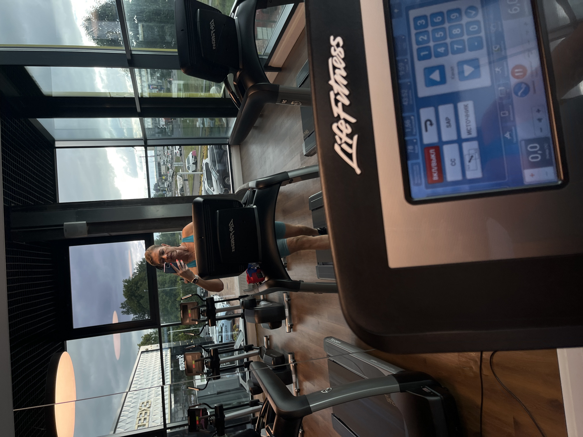 The image depicts the interior of a modern gym, likely located in Kaliningrad, Russia, as suggested by the Cyrillic text on the equipment. A woman with blonde hair, wearing a turquoise tank top and blue athletic shorts, is taking a selfie with her smartphone while standing on a Life Fitness treadmill. She is positioned in the center of the frame, with other treadmills and gym equipment visible around her. The gym features large windows offering a view of an urban landscape with a street, cars, and buildings. The overall scene conveys a sense of active lifestyle and contemporary fitness facilities.
