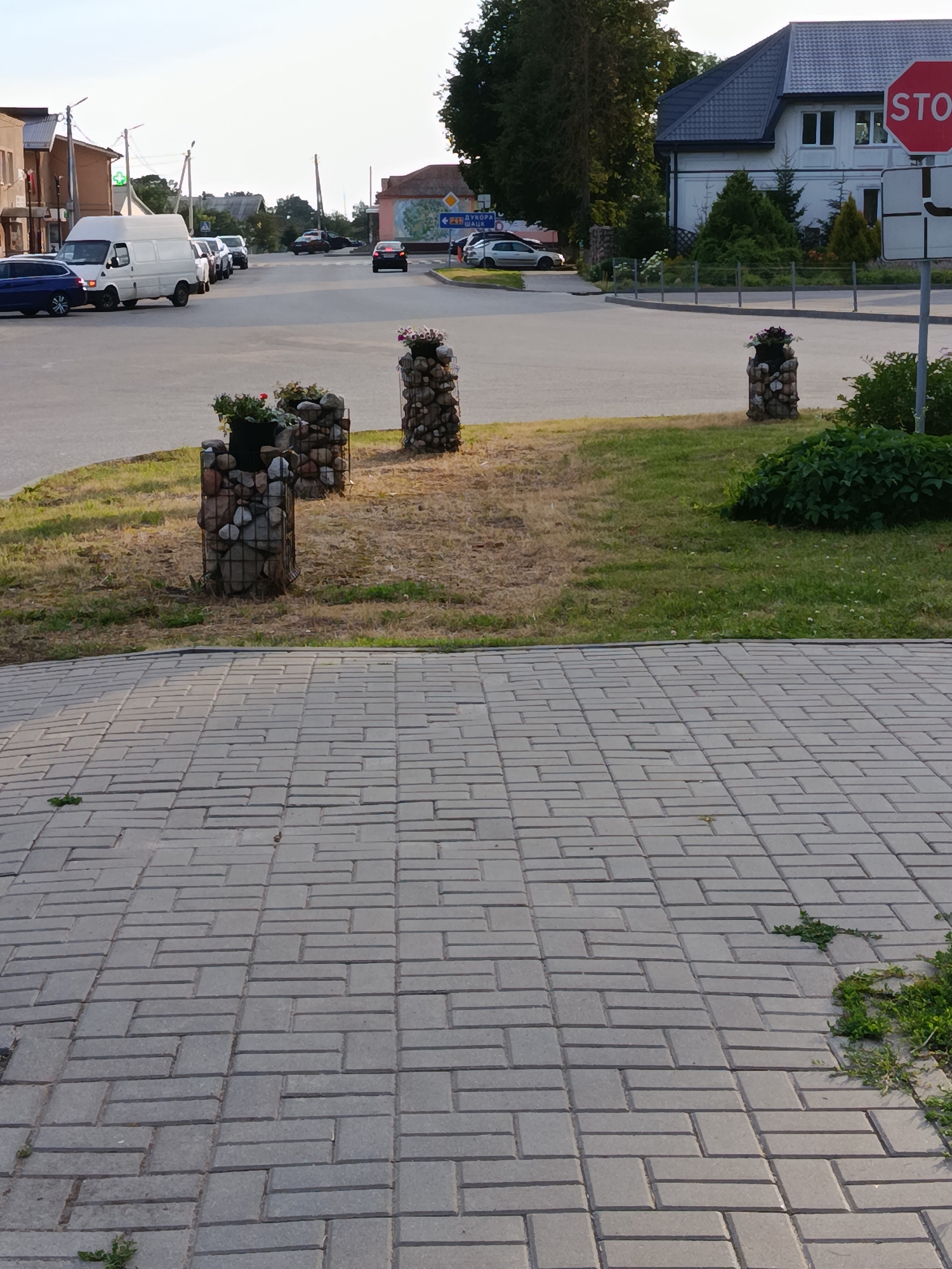 This media file captures a street scene in Smilavicki Selski Savet, Belarus. The foreground is dominated by a gray brick paved walkway, with patches of sparse grass and weeds growing between the bricks. In the middle ground, a well-maintained lawn area features three decorative planters. Each planter is a cylindrical wire cage filled with stones, topped with a dark pot containing small flowering plants and greenery. Beyond the lawn, a paved road curves into the distance, with several vehicles visible. A white van is stopped at an intersection, and other cars are seen in traffic.
On the right side of the frame, a modern white house with a dark roof stands prominently. Next to it, a red octagonal STOP sign is mounted on a pole. The background shows more buildings and trees, suggesting a semi-urban or suburban environment. The lighting indicates it is likely late afternoon or early evening, with soft shadows cast across the scene. The weather appears clear and sunny.
Visible text includes "STOP" on the sign. Additionally, a blue street sign is partially visible in the distance with text that appears to be in a Slavic language, possibly Belarusian or Russian, with the words "ДУКОВА" and "ШАЦК" discernible. No people are clearly visible in the image.