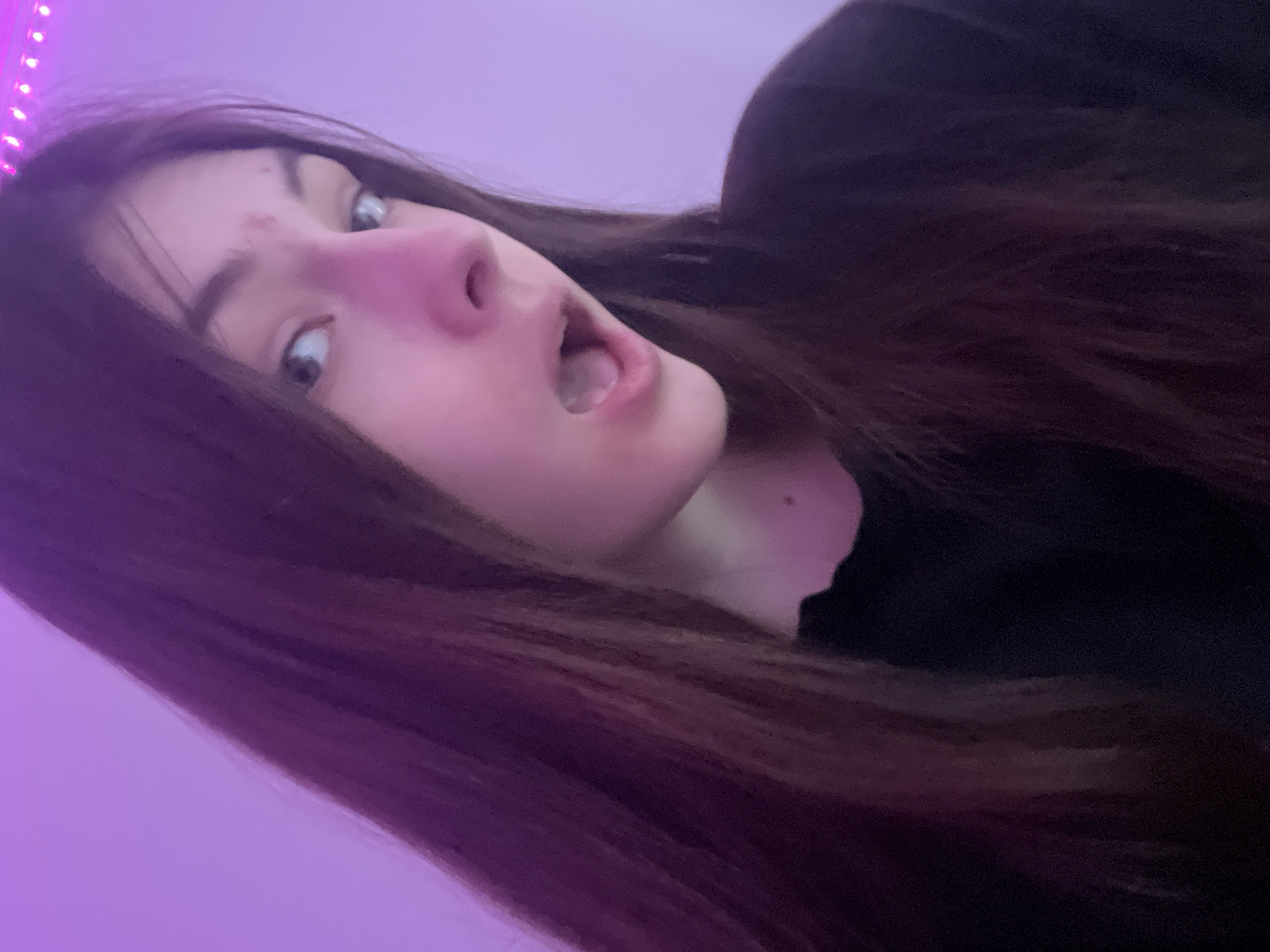 The media file presents a close-up, angled portrait of a young person, likely female, in an indoor setting, potentially in Saint Petersburg, Russia. The individual's face is dramatically tilted, creating a dynamic perspective as if looking directly up at the camera. She has long, dark brown hair that cascades around her face and shoulder.
Her expression is one of apparent surprise or playful shock, characterized by wide, light-colored eyes staring intently forward and an open mouth revealing some teeth and the inside. Her nose appears distinctly reddish-pink, possibly accentuated by the lighting or a natural flush. Small blemishes are visible on her forehead.
The background is dominated by a vibrant purplish-pink illumination, suggesting artificial light sources such as LED strips, with individual light points visible along the left edge of the frame. The person is wearing dark clothing, visible on the right side of the image. The setting appears to be a room, possibly a bedroom, creating an intimate atmosphere. The reliance on artificial light indicates it is likely evening or night. There is no visible text in the image.