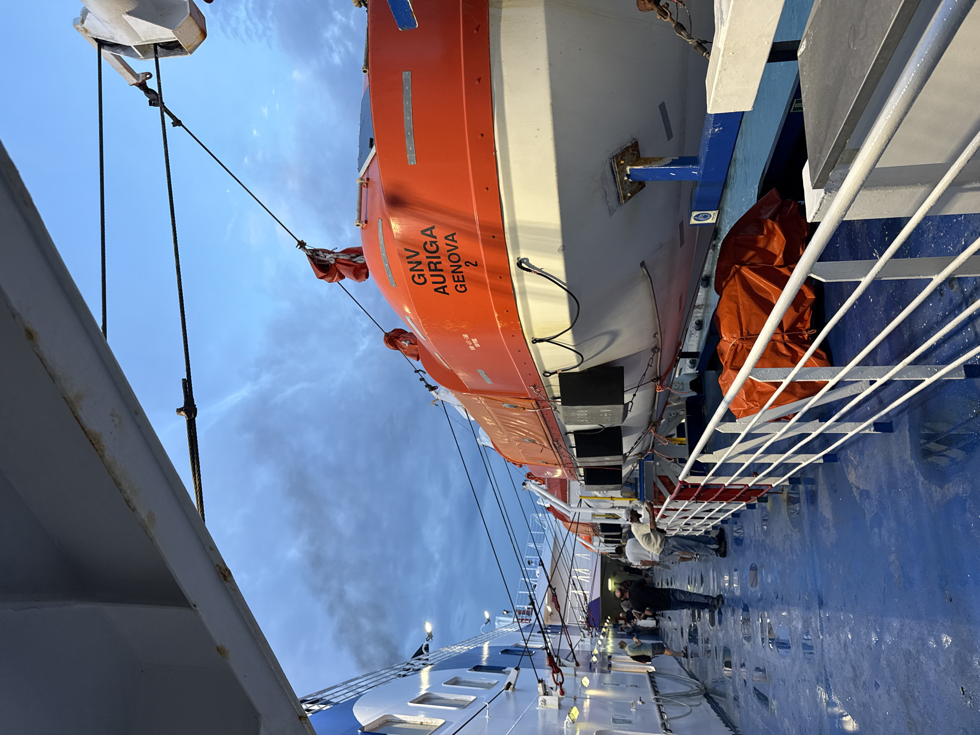 The image depicts the deck of a GNV (Grandi Navi Veloci) ferry, specifically the "AURIGA" bound for or docked in Genoa, Italy. A partially visible bright orange and white lifeboat marked "GNV AURIGA GENOVA 2" is attached to the side of the ferry. Several people are walking along the blue-painted deck. White railings are visible along the side of the ship. The sky is partially cloudy, suggesting dawn or dusk. The ferry appears to be docked next to another vessel. It is presumed the ferry is in Civitavecchia, Italy.