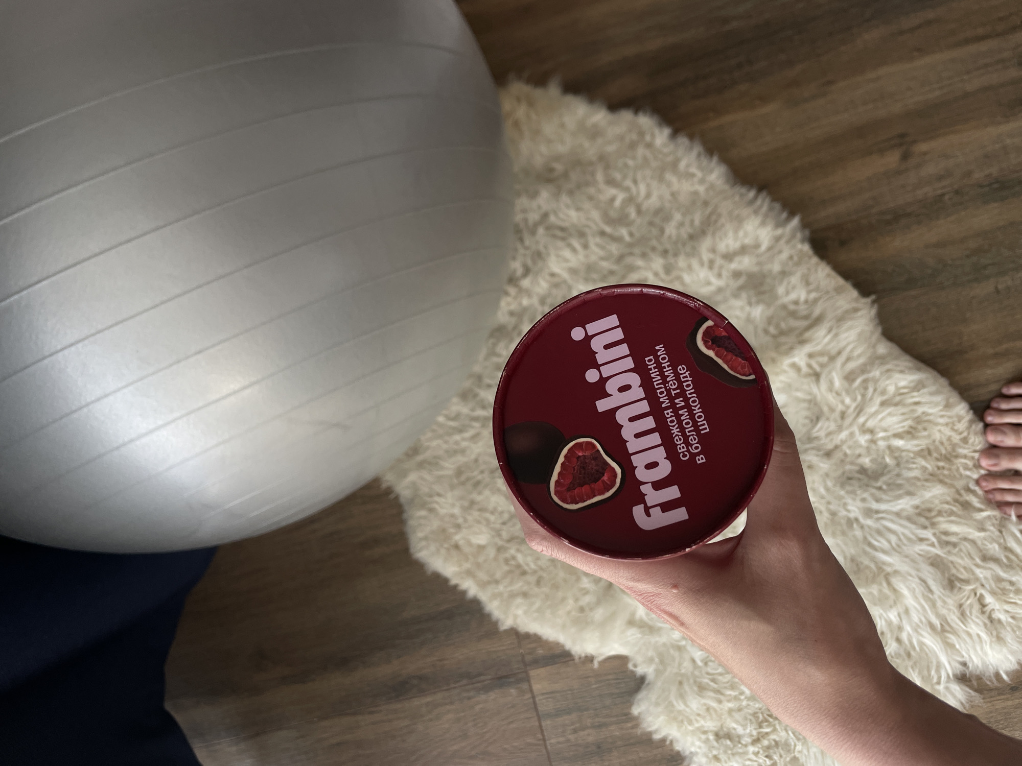 A photograph taken from an overhead perspective presents a scene indoors in Sepych, Russia. A person's hand holds a cylindrical container, the lid of which is predominantly maroon in color and labeled with a white inscription that reads "Frambini." The lid also features an image of a dark chocolate-covered raspberry.
Adjacent to the hand and the container is a plush, light-colored rug. In the background, a silver-colored exercise ball is partially visible. The floor is made of wooden planks, and the individual's toes are observable on the right side of the photograph. The person is wearing navy blue pants.