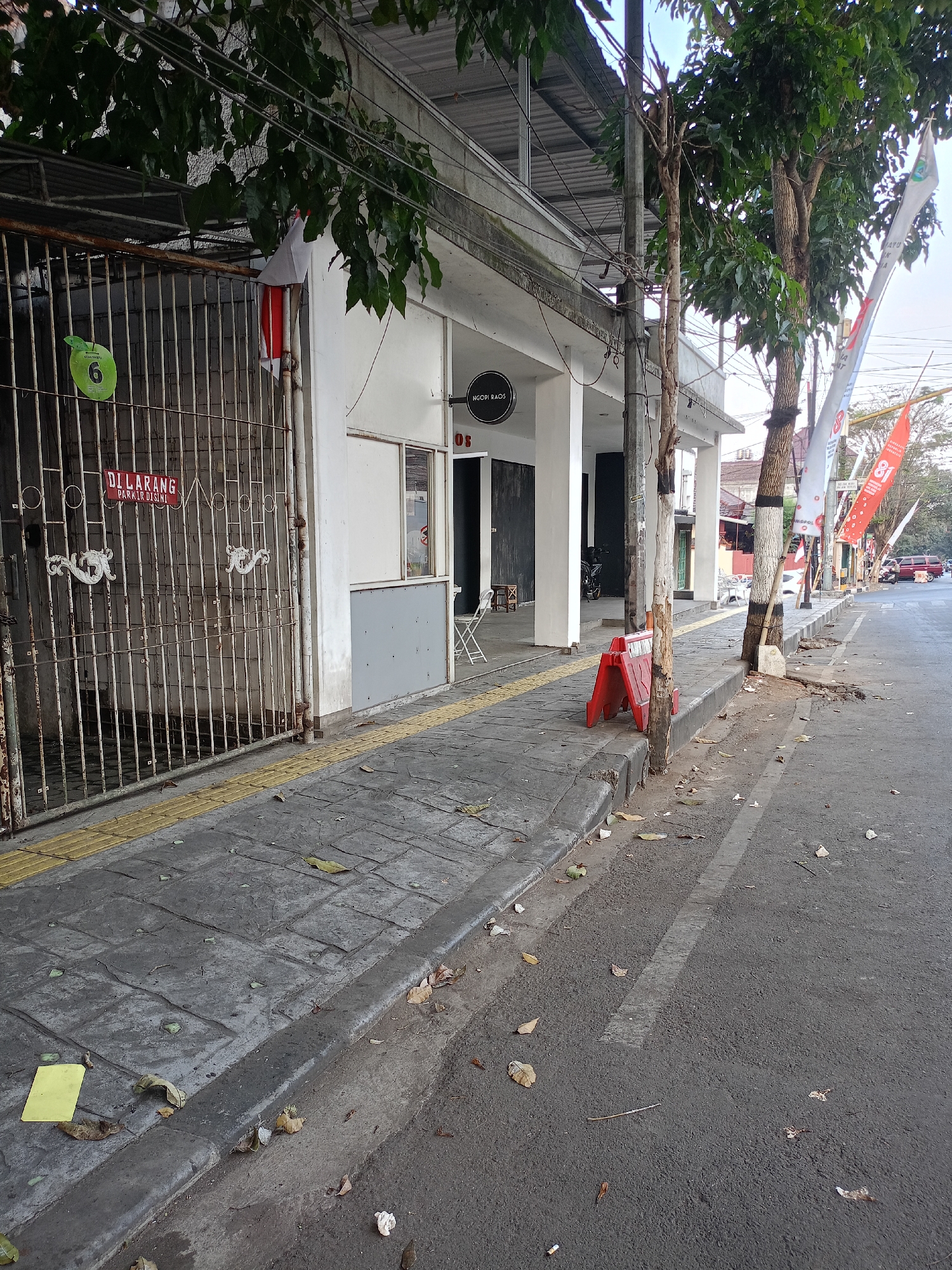 The image depicts a street scene in Batu, Indonesia, on a clear day. A sidewalk runs along the left side of the image, featuring textured paving stones and a yellow tactile paving strip. Adjacent to the sidewalk is a white-fenced gate with decorative scrollwork and a green sign with the number 6, and a red sign with the text "DILARANG PARKIR DI SINI" (No Parking Here). A white building with columns stands behind the gate. Further down the sidewalk, a red traffic barrier is placed near a tree. Beyond the building, the street is visible with several trees lining the right side, some adorned with flags. A few vehicles are present in the background. The ground on the sidewalk and street is scattered with fallen leaves. The overall impression is a street in a developed urban area.