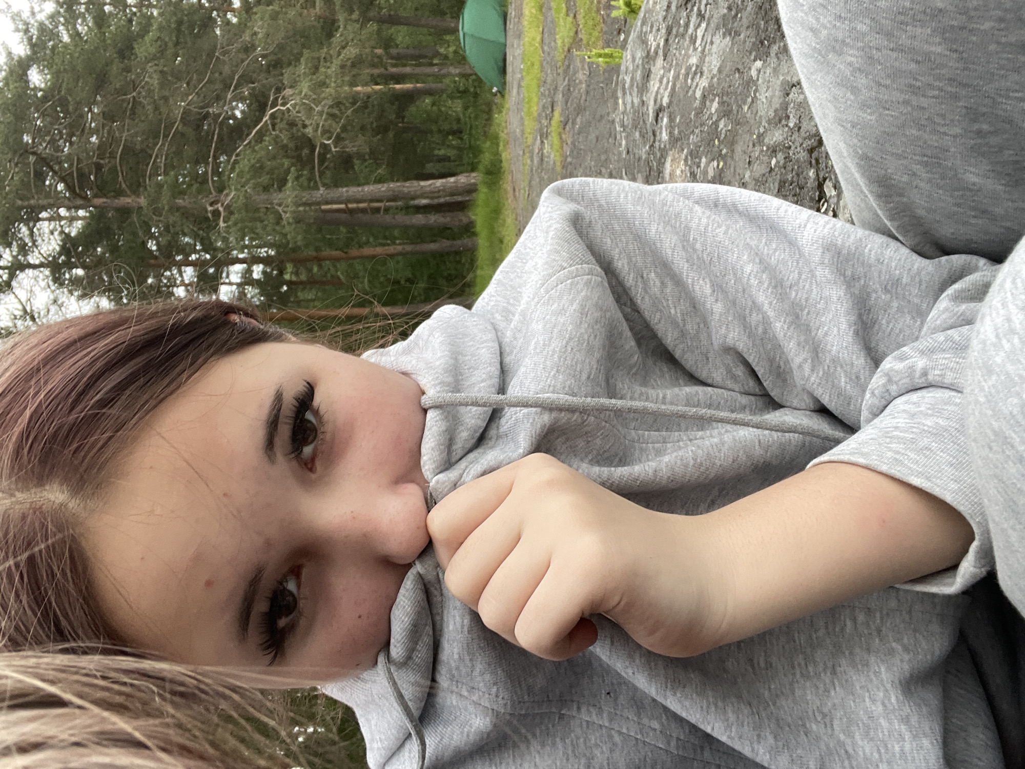 A young individual is captured in a close-up, selfie-style photograph, situated outdoors in what appears to be a natural, forested environment in Boitsovo, Russia. The individual, whose gender cannot be definitively determined from the visible features but presents as female, has dark hair partially obscuring the left side of their face (viewer's left) and visible dark eyes framed by prominent, long eyelashes. Small blemishes are present on the forehead and upper cheeks. They are wearing a heather grey hooded sweatshirt (hoodie) with a visible drawstring. Their right hand (viewer's left) is holding the collar of the hoodie up to obscure their mouth and nose. The individual's gaze is directed straight at the camera. The person appears to be seated on a large, flat, grey rock formation, which occupies the lower portion of the frame and shows patches of moss and sparse green vegetation. In the background, a dense forest of tall, slender evergreen trees with green canopies is visible. A patch of green grass lies between the rock and the tree line. A small, green dome tent is pitched on the grassy ground, positioned towards the right side of the background. The sky, partially visible through the trees, is overcast and uniformly light, indicating diffused natural light.