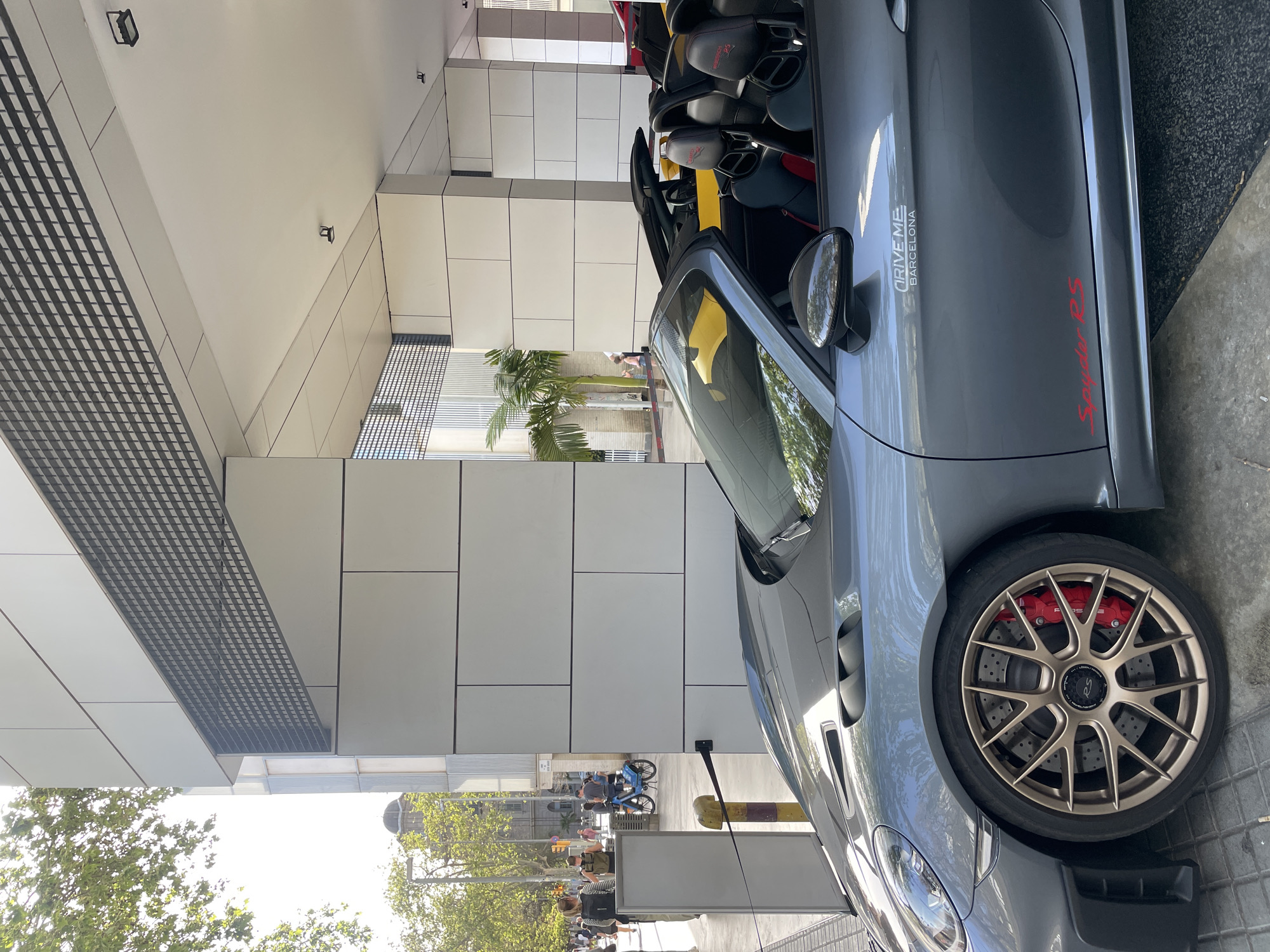 The image depicts a grey Porsche Spyder RS parked on a street in Barcelona, Spain. The car has bronze-colored wheels, red brake calipers, and a sticker on the side that says "DRIVE ME BARCELONA." The car's top is down, revealing yellow interior accents and black seats.
In the background, there is a modern building with a distinctive facade composed of white panels and a metallic, grid-like design on the overhang. Further into the background, there are trees, a street with pedestrians, and a cyclist on a blue bicycle. The overall lighting suggests it is daytime.