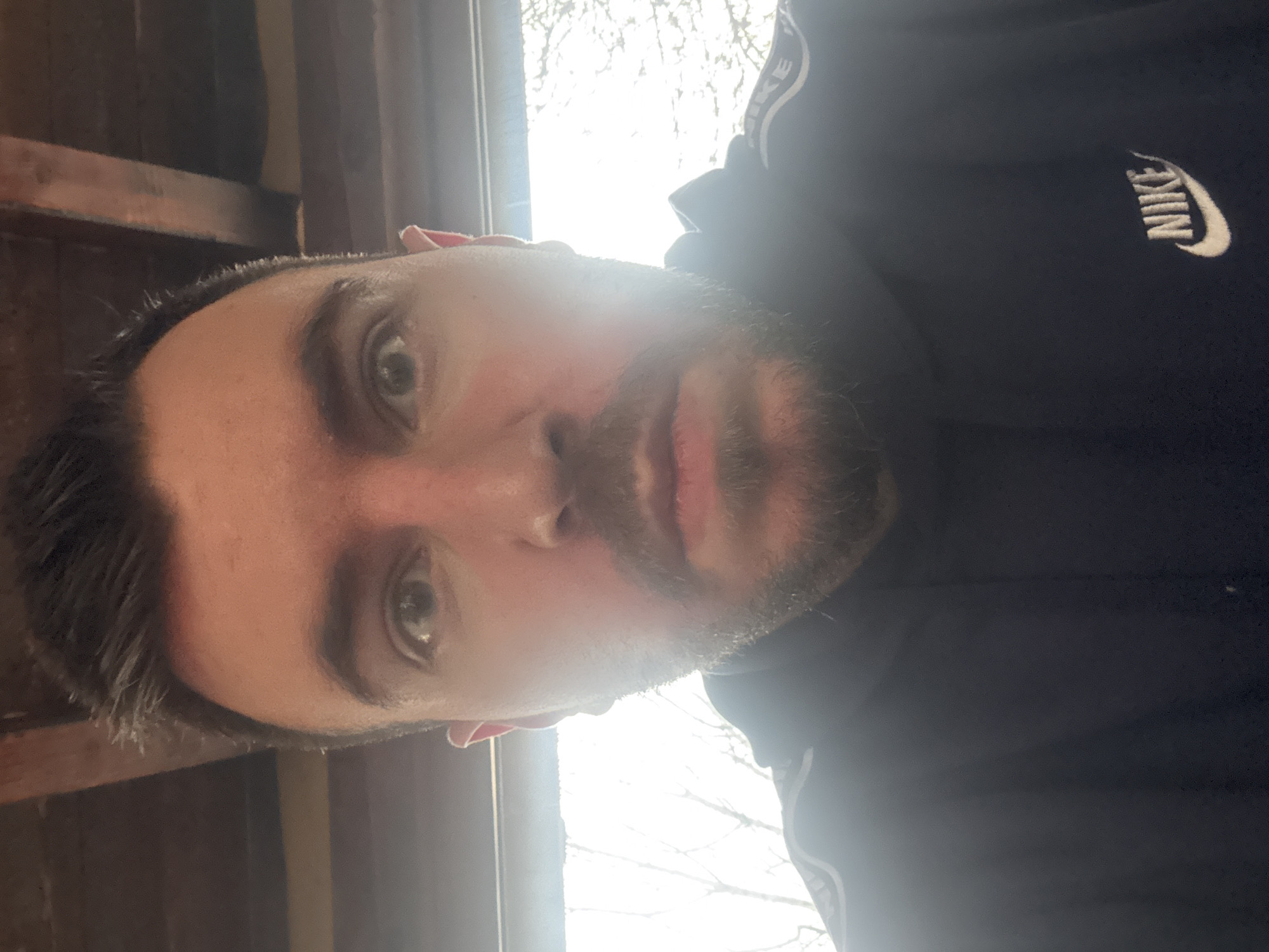 A close-up selfie of a man with dark hair and a beard, wearing a black Nike jacket, is visible. The photo is taken indoors, near a window. The background suggests a room with wooden elements, possibly a part of the ceiling or wall. The bright daylight coming through the window washes out some details. There's no specific indication of the location being Sofia, Bulgaria, nor can any information about actions or interactions be determined.
