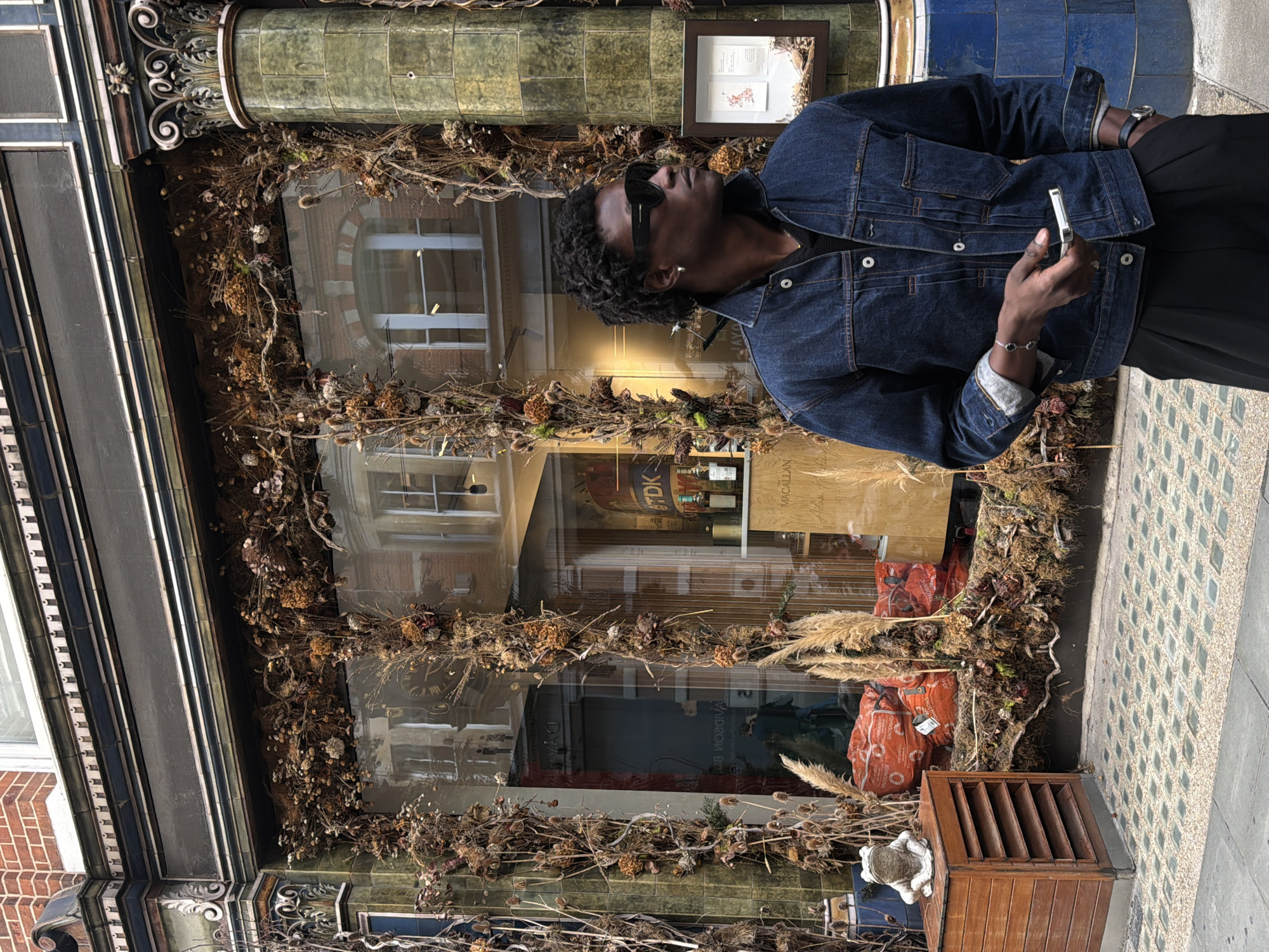 A man stands on a sidewalk in front of a shop window, which is decorated with dried flowers and branches. The man is wearing a denim jacket, black pants, sunglasses, and a watch. He is holding a phone in his right hand. The shop window reflects buildings across the street. The sidewalk is paved with cobblestones and grey concrete. The shop has a brick facade and decorative columns. A wooden cabinet with a frog statue sits to the left of the shop. The scene appears to be in London, United Kingdom, based on the architectural style and the presence of a red telephone box visible in the reflection.