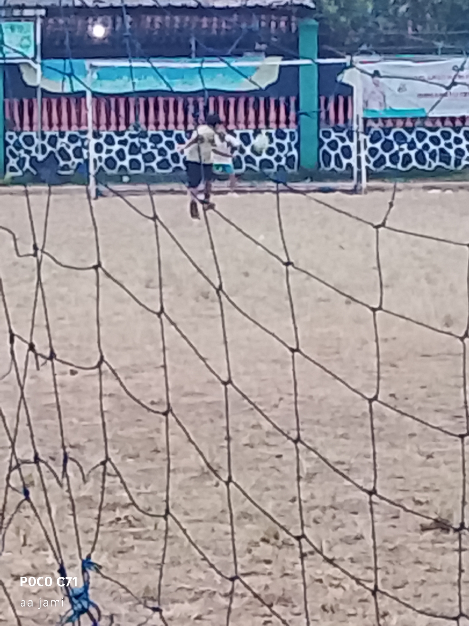 The image depicts a low-angle shot of a dirt field, likely a sports ground, with a black net in the foreground obscuring parts of the scene. In the background, two individuals, appearing to be young males, are engaged in an activity. One individual, wearing a light-colored shirt and dark shorts, is in motion, possibly kicking or interacting with a ball, though the ball is not clearly visible. The other individual, similarly dressed, is positioned nearby. Behind them is a fence with a decorative pattern of black shapes on a white background, topped with a green structure and a pink and white striped railing. A banner is visible on the right side of the fence. The environment appears to be outdoors with some greenery and overcast sky suggested by the lighting. The watermark "POCO C71 aa jami" is present in the bottom left corner of the image. The scene is consistent with a local sports field in Tasikmalaya, Indonesia, where young people often gather for recreation.