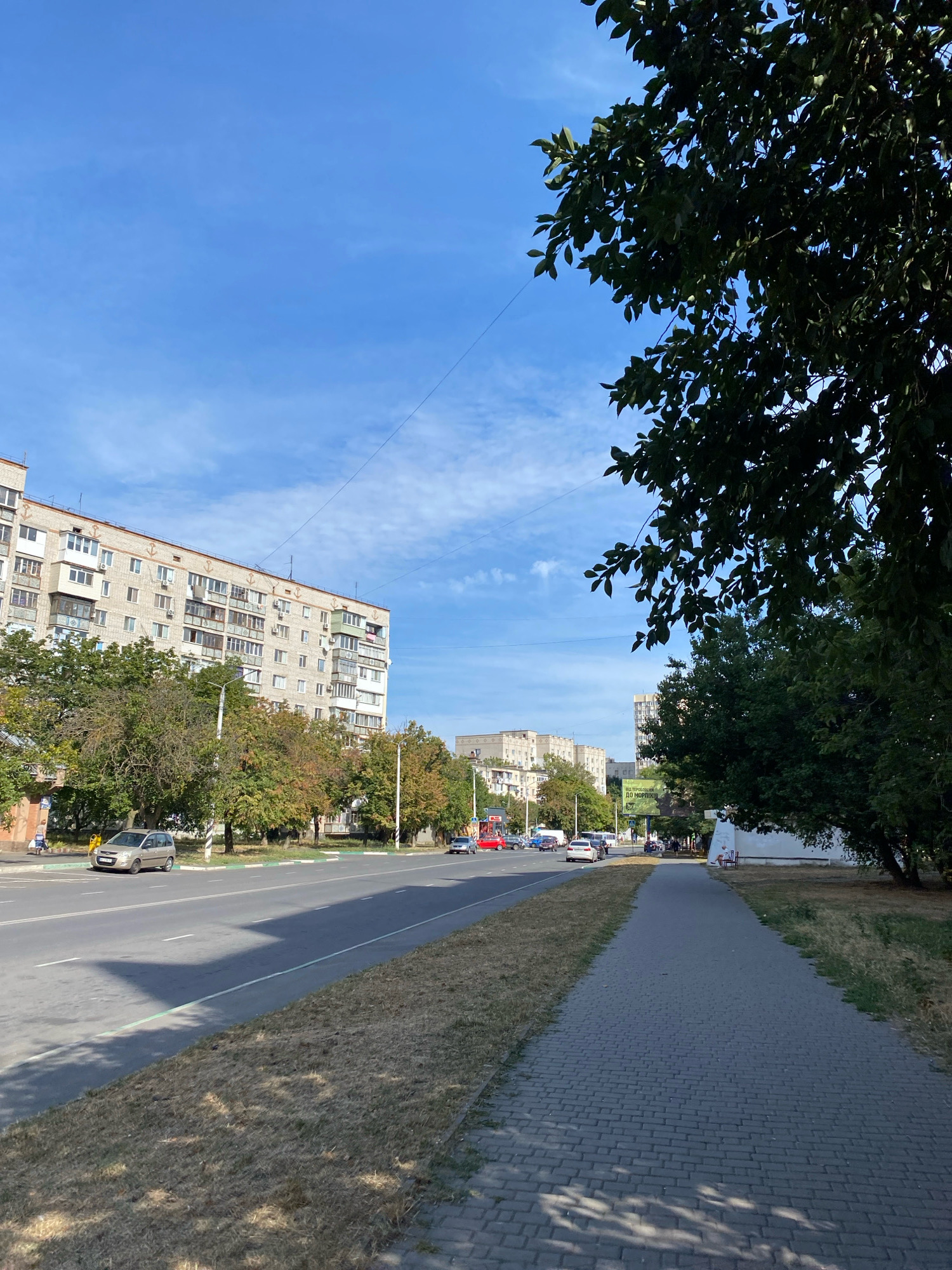 The image captures a street scene in Chornomorsk, Ukraine, under a bright blue sky with sparse white clouds. A large, multi-story apartment building with numerous balconies dominates the left side of the frame, featuring light-colored facades and decorative anchors along its upper edge. A row of trees with green and autumnal foliage lines the sidewalk and separates the street from the building.
The street itself is paved and appears to be a multi-lane road. Several vehicles are visible, including a silver hatchback parked on the side, and cars driving on the road. A paved pedestrian walkway runs along the right side of the street, bordered by dry grass and trees. In the background, more apartment buildings are visible, along with streetlights and some signage, including a billboard. The overall atmosphere suggests a typical urban environment on a sunny day.