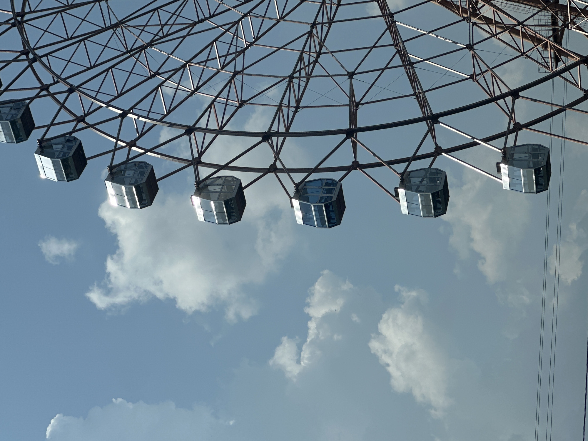 The image is a close-up, low-angle shot of a Ferris wheel against a partly cloudy blue sky. Several gondolas of the Ferris wheel are visible, appearing as enclosed, boxy cabins with reflective glass windows. The metal structure of the Ferris wheel is intricate, with a dark, reddish-brown frame. In the distance, power lines are visible. The overall impression is of a bright, sunny day at an amusement park or fairground. The Ferris wheel is likely the "Chum" Ferris wheel located in Novosibirsk, Russia, due to its distinctive design and common association with city skylines in Russia. However, no specific landmarks confirm this location from the provided image. There are no people visible in the image.