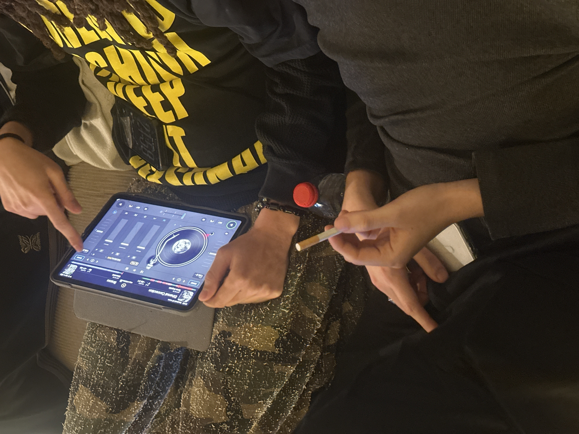 A close-up, overhead view depicts three individuals in an indoor setting. One individual, positioned on the left and center, operates a tablet identified as an iPad, encased in a light grey folio, displaying a DJ mixing application featuring virtual turntables, audio waveforms, and the track "Ethereal Connection." Another individual's hand, entering from the upper frame, points at the tablet screen. A third individual, partially visible on the right, wears a black hoodie adorned with yellow striped text and possesses dark dreadlocks. This individual holds an unlit cigarette in their right hand and a clear plastic bottle with a red screw cap in their left hand. A white smartphone is partially visible, tucked into their lap. A black beaded bracelet is worn on the left wrist of this individual. The immediate environment consists of various soft textiles and fabrics, consistent with a casual, relaxed indoor atmosphere. The specific location within the city of Paris, France, cannot be determined from the visual information provided.